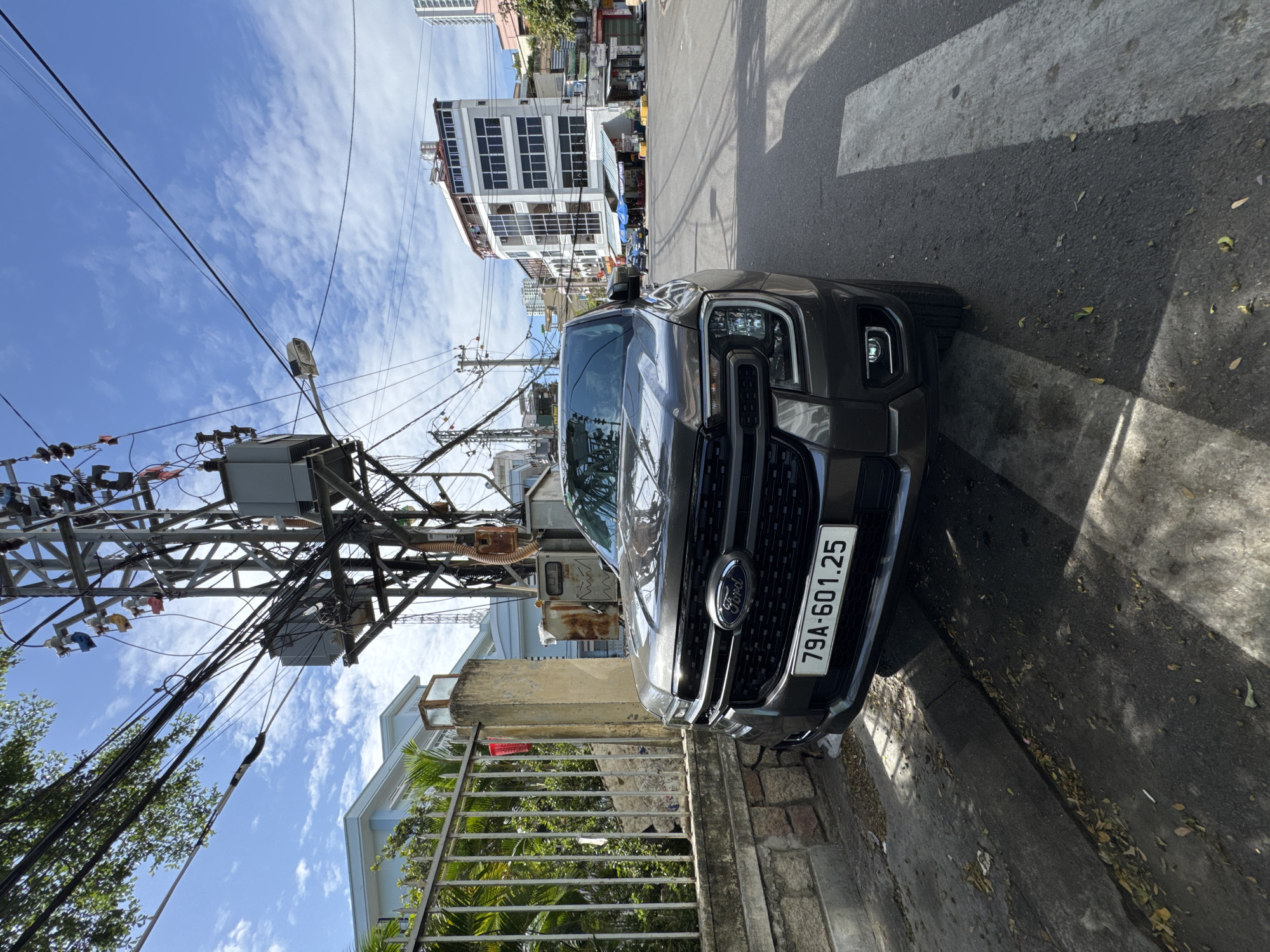 In Nha Trang, Vietnam, a dark gray Ford Ranger pickup truck is parked on the right side of a paved street. The truck is facing slightly towards the viewer, with its front grille and headlights clearly visible. A license plate with the characters "79A-601.25" is affixed to the front of the vehicle.
To the left of the truck, a low stone wall topped with a metal fence runs along the sidewalk. Beyond the fence, palm trees and other greenery are visible. A concrete pillar stands next to the fence, with a small red object hanging from it.
The street is lined with buildings, including a modern, multi-story white building with many windows to the right of the truck. Overhead, a complex network of electrical wires and poles dominates the sky, with transformers and various insulators attached. The sky is blue with scattered white clouds. Sunlight casts shadows on the street and sidewalk, indicating it is daytime. A pedestrian crossing is visible on the street in front of the truck. The overall impression is an urban street scene in Vietnam.