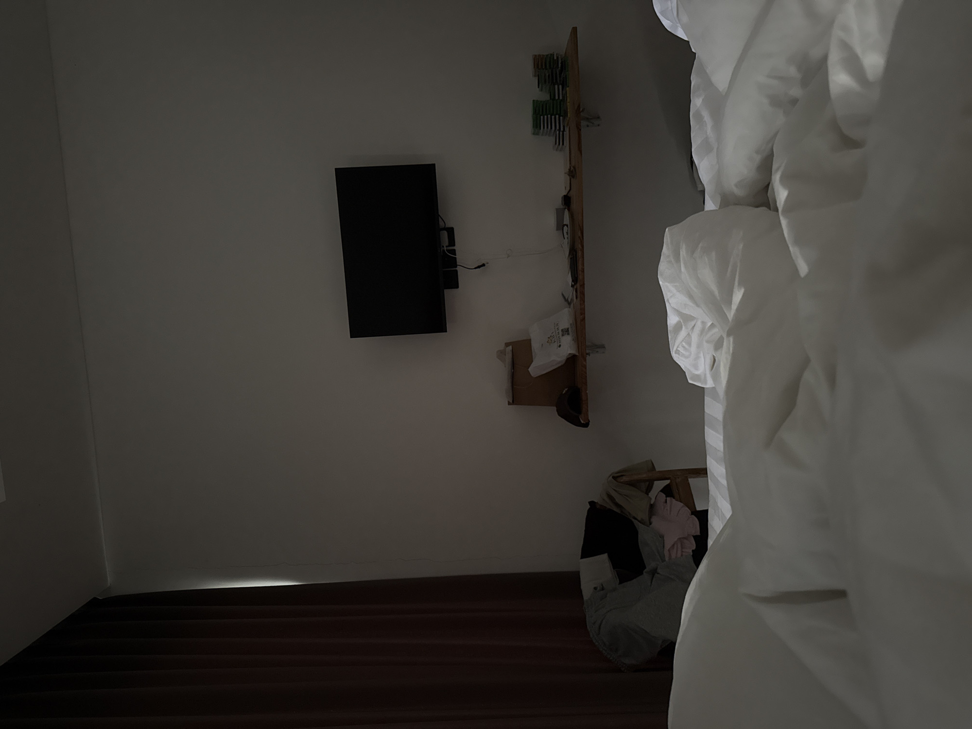 The image depicts a dimly lit hotel room in North Kuta, Indonesia. In the foreground, a rumpled bed with white linens dominates the frame. To the left, a dark brown curtain is partially drawn, allowing a sliver of light to illuminate a crack in the wall. Further back, a wooden chair is draped with assorted clothing, including pants, a shirt, and what appears to be a pink garment. On the wall above, a black flat-screen television is mounted, with a black bracket and cables visible beneath it. A wooden shelf is fixed to the wall below the television, holding various items, including a white bag, what looks like a hat, and several stacks of what could be video game cartridges or coasters. The environment appears to be a domestic interior, likely a hotel room given the presence of a television and the overall neatness, despite the scattered clothing.
