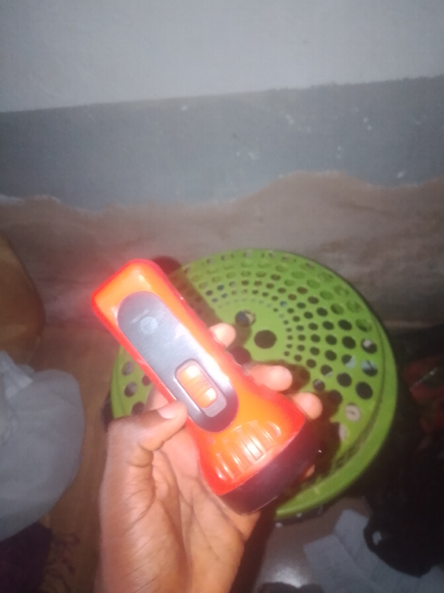 A close-up, handheld shot shows a person's left hand holding a red and gray flashlight. The flashlight is positioned in the foreground, obscuring much of the background. The person's skin tone is dark, and their fingers are wrapped around the body of the flashlight. The flashlight has a prominent orange button and a gray section with some text and markings. Behind the flashlight, a bright green, circular object with a perforated pattern is visible. The background consists of a mottled, light-colored surface that appears to be a wall, with some indistinct dark shapes and areas of shadow. The overall lighting is dim, and the image quality is somewhat blurry. The location is stated to be Jalingo, Nigeria.