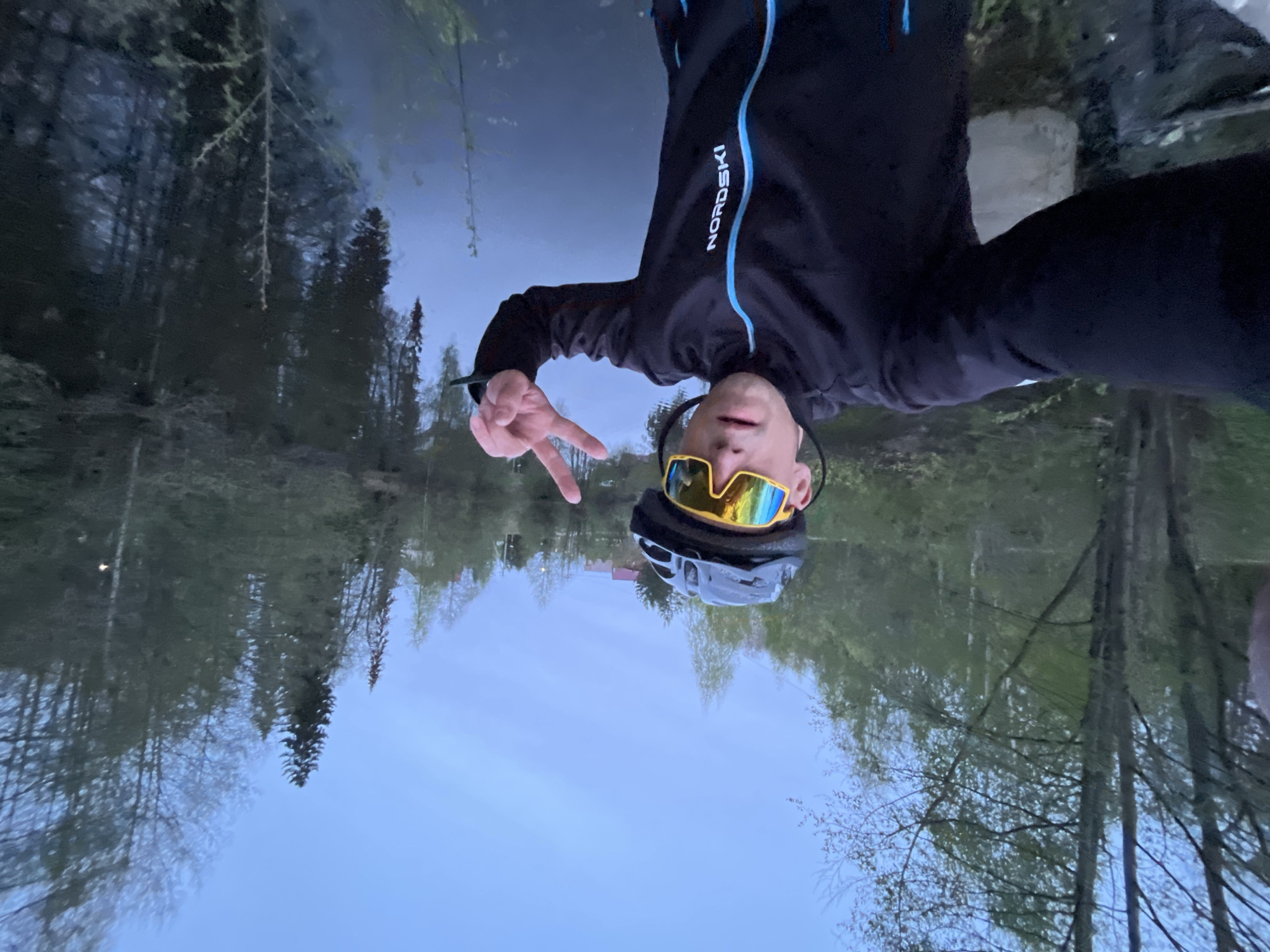 The image depicts a person wearing a black "Nordski" jacket with blue trim, a helmet, and yellow/blue mirrored sunglasses. They are holding up a peace sign with their hand. The background appears to be a body of water, likely a lake or river, reflecting a treeline and the sky. It is dusk or dawn, based on the lighting. Given the context of "Chaikovsky, Russia", this could be a lake or river in or near the city. More specifically, it is the Kama Reservoir in Chaikovsky, Russia.