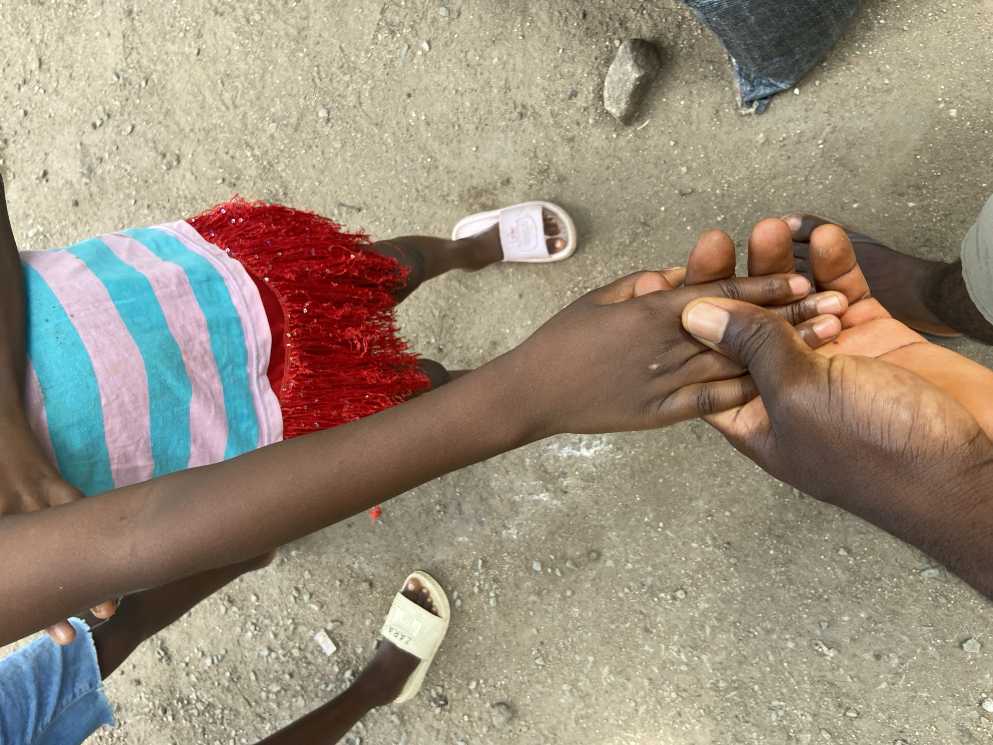 The image depicts a top-down view of two individuals, likely in Mararaba, Nigeria. One individual, possibly a child, is wearing a striped shirt with red fringe, blue shorts, and two different colored slippers: one white and one pink. The other individual is holding the first individual's hand. The background shows a concrete surface with scattered pebbles and a rock. A dark gray bag is also visible in the upper right corner of the image.