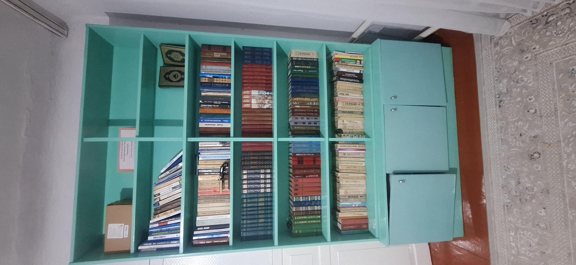 The image depicts a turquoise bookshelf filled with numerous books. The bookshelf is situated in what appears to be a room with plain white walls and a light-colored patterned carpet on the floor. On the top shelf, to the left, there is a brown cardboard box with a label. To its right, a pinkish certificate or sign is affixed to the shelf. The middle shelves are densely packed with books of various sizes and colors, some standing upright and others leaning. On the second shelf from the top, in the right section, two ornate, dark-colored books are placed side-by-side. The bottom section of the bookshelf consists of two cabinet doors with silver handles, partially obscuring the contents within. The floor beneath the bookshelf is a reddish-brown color. The overall environment suggests an indoor setting, possibly a home or a small library, located in the city of Novopokrovka, Kyrgyzstan. No people are visible in the image.