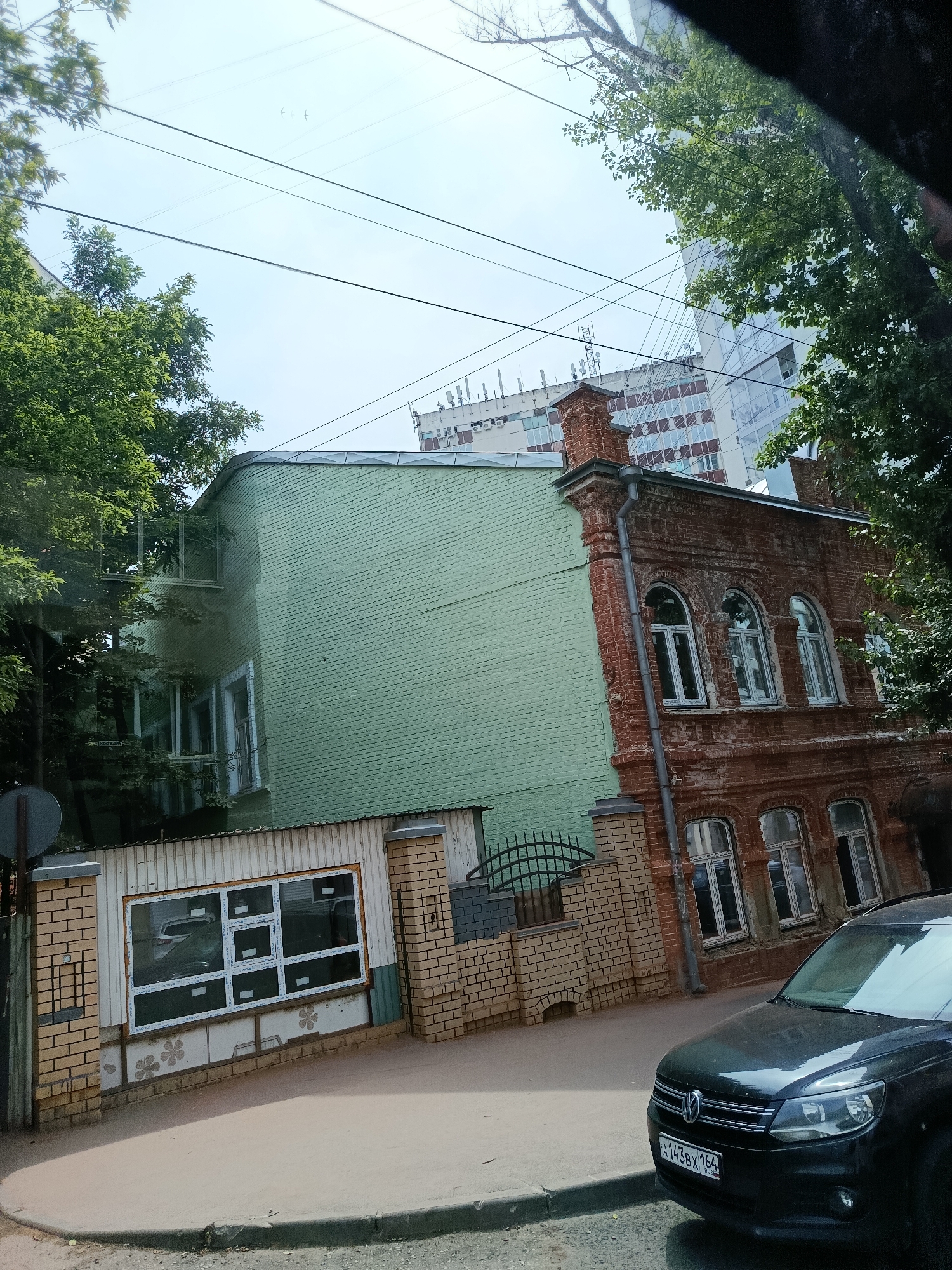 The image depicts a street scene in Saratov, Russia, showcasing a juxtaposition of old and new architecture. On the right, a weathered, red brick building with arched windows, characteristic of older construction, stands prominently. Adjacent to it, on the left, is a more modern, light green painted brick building. The green building appears to have been built or renovated in a way that partially obscures the older structure. In the foreground, a dark-colored Volkswagen SUV is parked. The street is paved with asphalt and has a curb. A white metal fence with brick pillars runs along the property line. In the background, a modern, multi-story building with a reflective glass facade rises above the older structures, indicating urban development. Trees with lush green foliage are scattered throughout the scene, partially obscuring the sky and the upper portions of the buildings. The sky is visible with some power lines crossing it, and two birds can be seen flying in the upper portion of the image. The overall impression is of a street in a Russian city undergoing modernization while retaining elements of its historical past.
