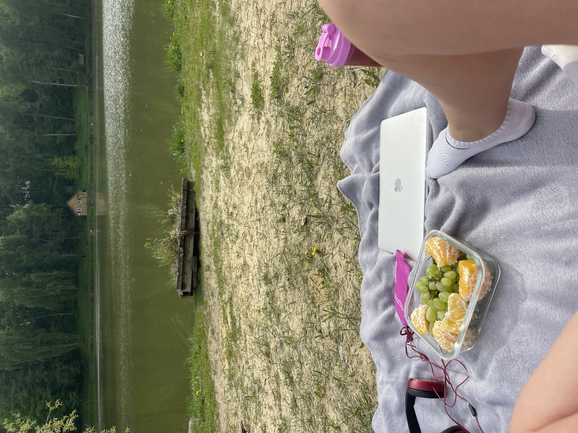 The image captures a scene at a lake in Misaylovo, Russia. The foreground features a gray blanket spread on a sandy area with sparse vegetation. On the blanket, there's a silver laptop, a clear glass container with orange segments and grapes, and a pair of red headphones. A pink water bottle is also visible. The scene includes a person's legs and feet wearing white socks. The lake is in the background, with a wooden structure near the edge and trees lining the opposite shore. A small building is visible in the distance across the lake.