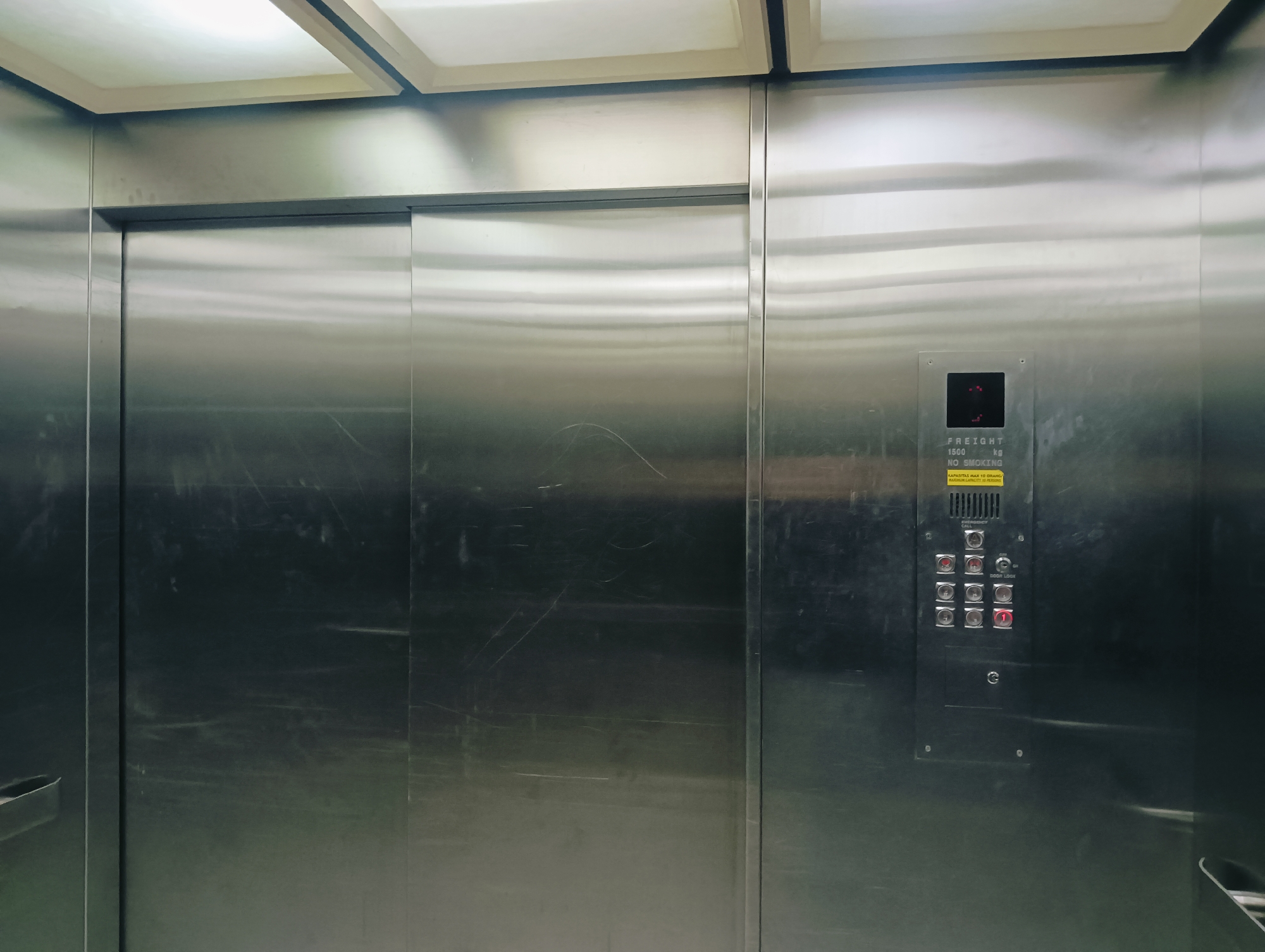 The image shows the interior of a stainless steel elevator. The elevator doors are closed and appear to be sliding doors. On the right side of the elevator wall, there is a control panel with buttons and a digital display. The display shows a red "1". Above the display, there is text indicating "FREIGHT 1500 kg NO SMOKING". Below the digital display, there is a panel with several red buttons, some of which are illuminated. There is also a yellow sticker with text that is partially obscured by a button but appears to be related to capacity. The ceiling of the elevator is illuminated by fluorescent lights. The walls of the elevator are scratched and smudged, indicating heavy use. Based on the text on the control panel, this elevator is located in Batam, Indonesia.