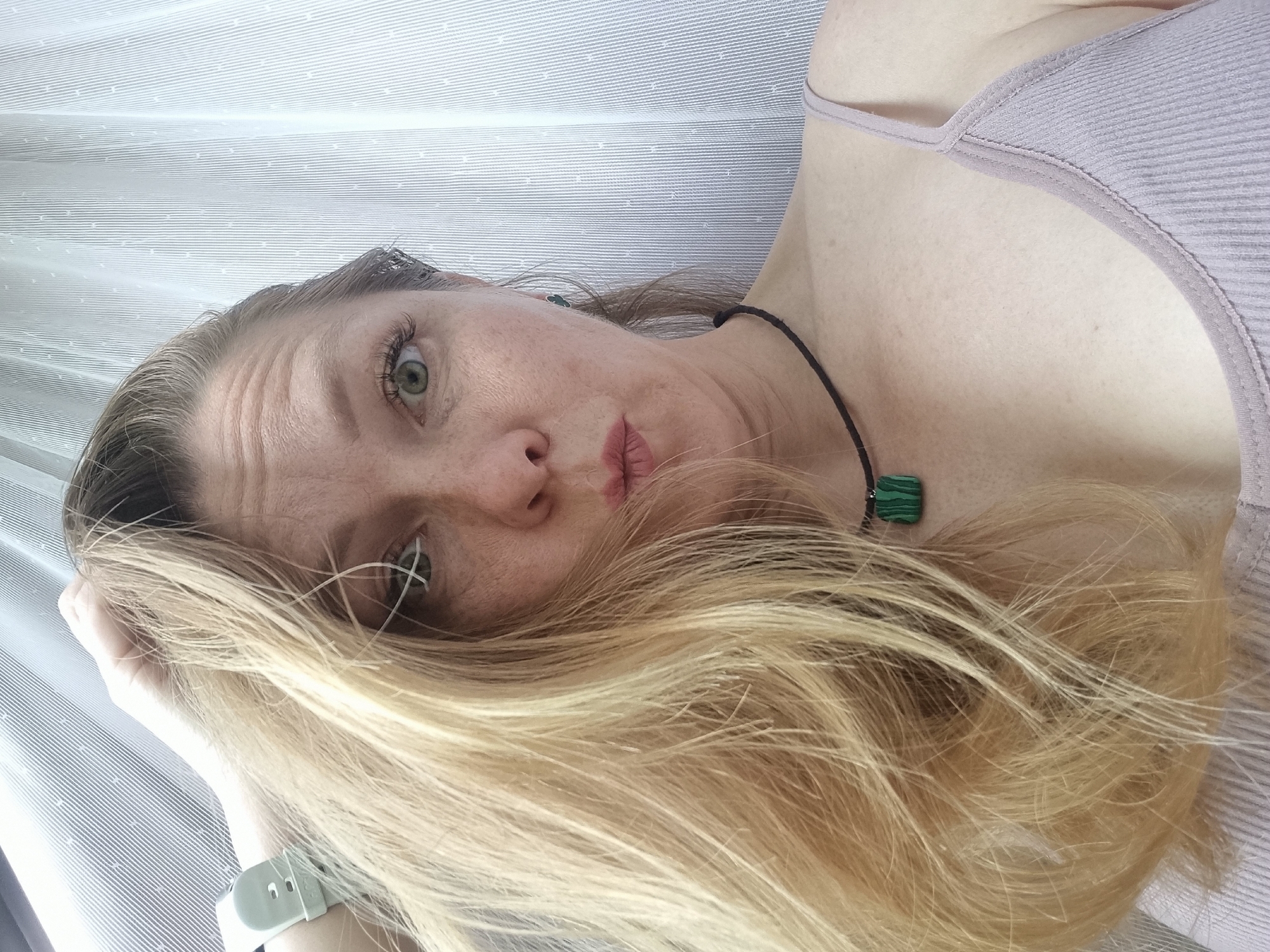 A woman is taking a selfie indoors, possibly in her home in Chaikovsky, Russia. She has blonde hair that is falling over her face, obscuring part of her features. She is wearing a light-colored tank top and a necklace with a green pendant. Her hand is raised to her head, and she appears to be making a puckered-lip expression. The background is a white curtain with a subtle vertical pattern.