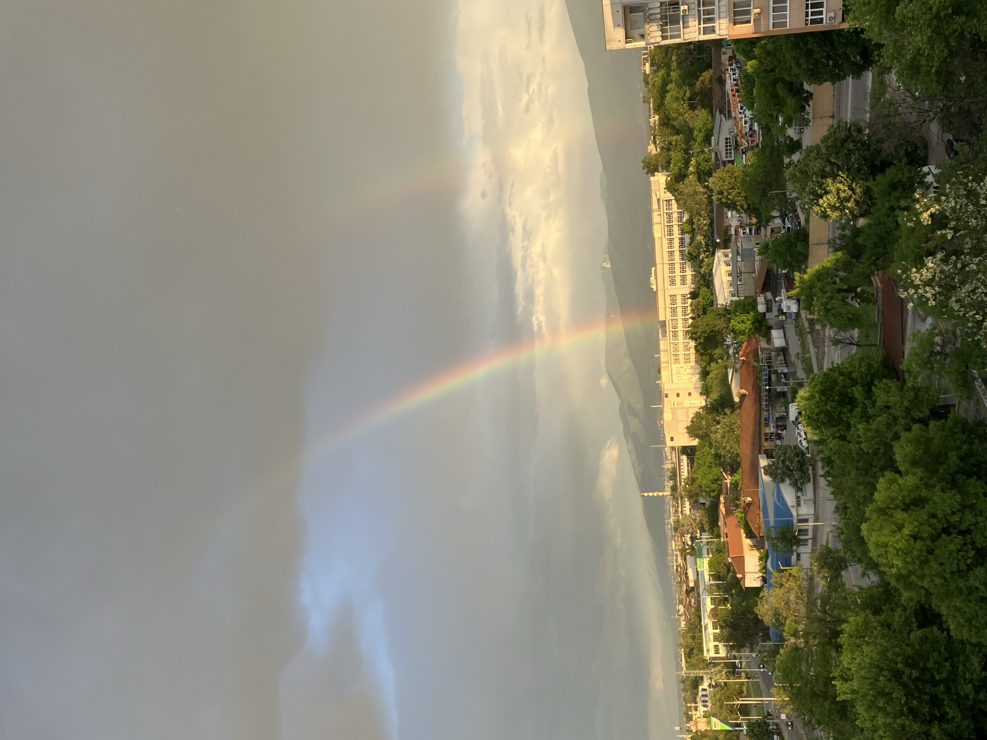 The image shows a view of the city of Plovdiv, Bulgaria, with a rainbow visible in the sky. The rainbow stretches across the sky from left to right, ending near a tall, white building in the city. The foreground shows trees, buildings, and streets. The background features the cloudy sky and mountains. The shot is taken from an elevated position, likely a high-rise building.