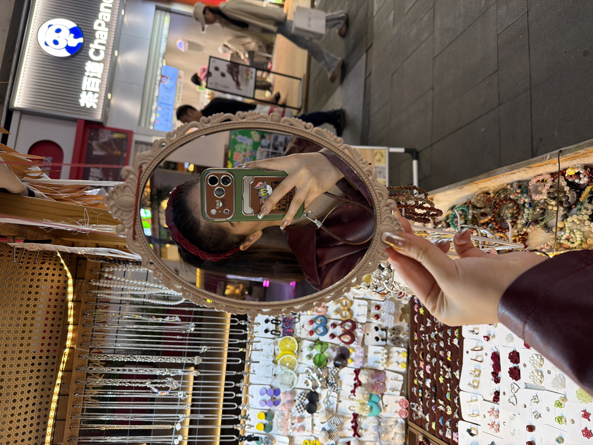 A person holds an ornate, oval mirror in front of a display of costume jewelry, capturing a selfie. The mirror reflects the person's face, wearing a red headband and holding a phone with a cartoon-themed case. The background, visible in the mirror, includes a street scene with pedestrians and a store sign in Chinese, partially identifiable as "ChaPanda." The jewelry display consists of multiple racks filled with necklaces, earrings, and other accessories. The person's arm and hand, clad in a maroon jacket, are visible holding the mirror. The image appears to be taken in a commercial area in the city of Chongqing High-tech Zone, China.
