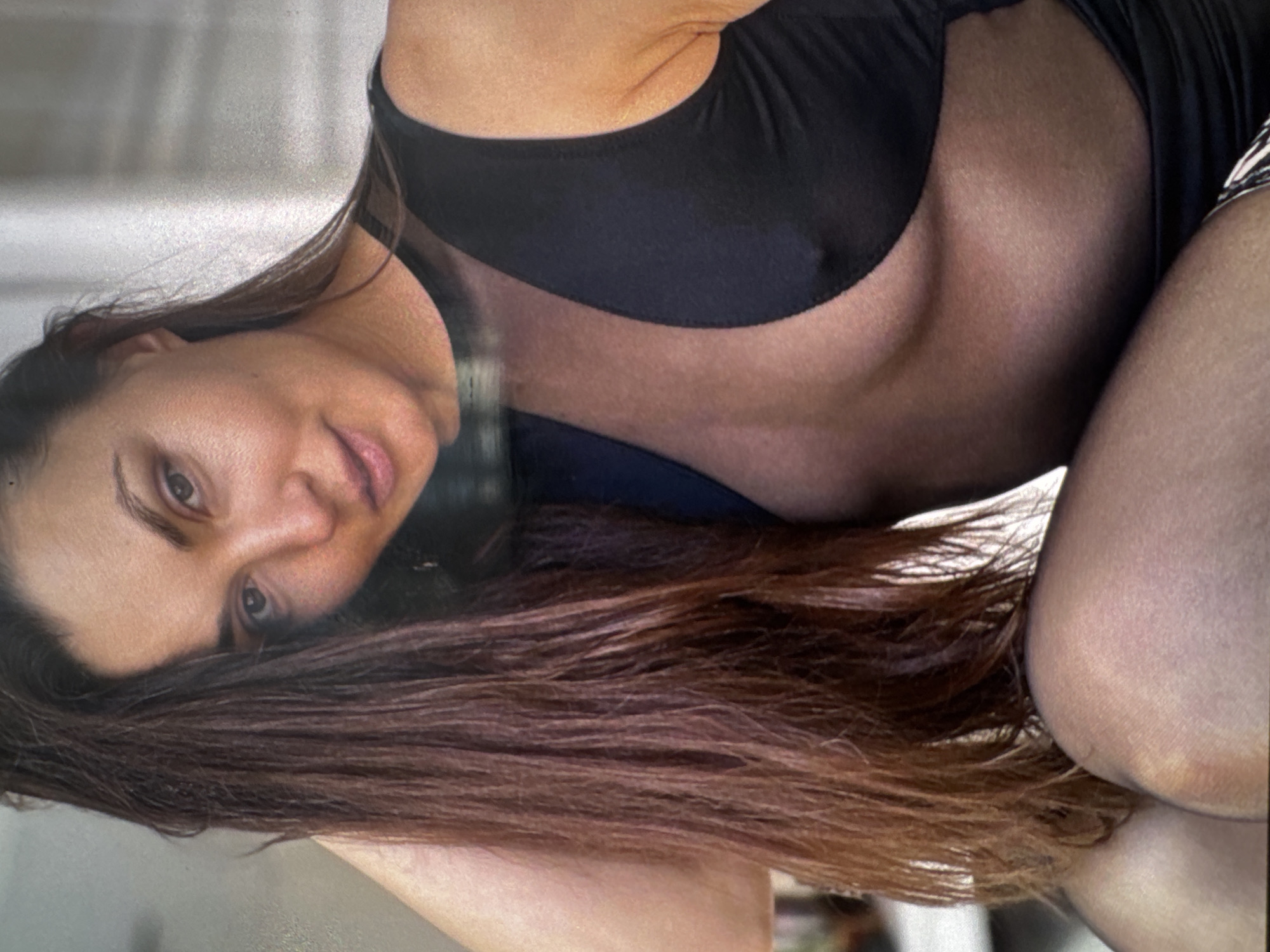 A close-up, waist-up shot shows a woman with long, dark brown hair in Sofiivska Borshchahivka, Ukraine. She is looking towards the camera with a neutral expression and her left arm is bent and resting behind her head. She is wearing a black, sleeveless dress with a sheer neckline that reveals her chest and shoulders. The dress has a visible seam running down the center of her chest. Her skin appears to be lightly tanned. The background is blurred and appears to be an indoor setting with a light-colored wall.