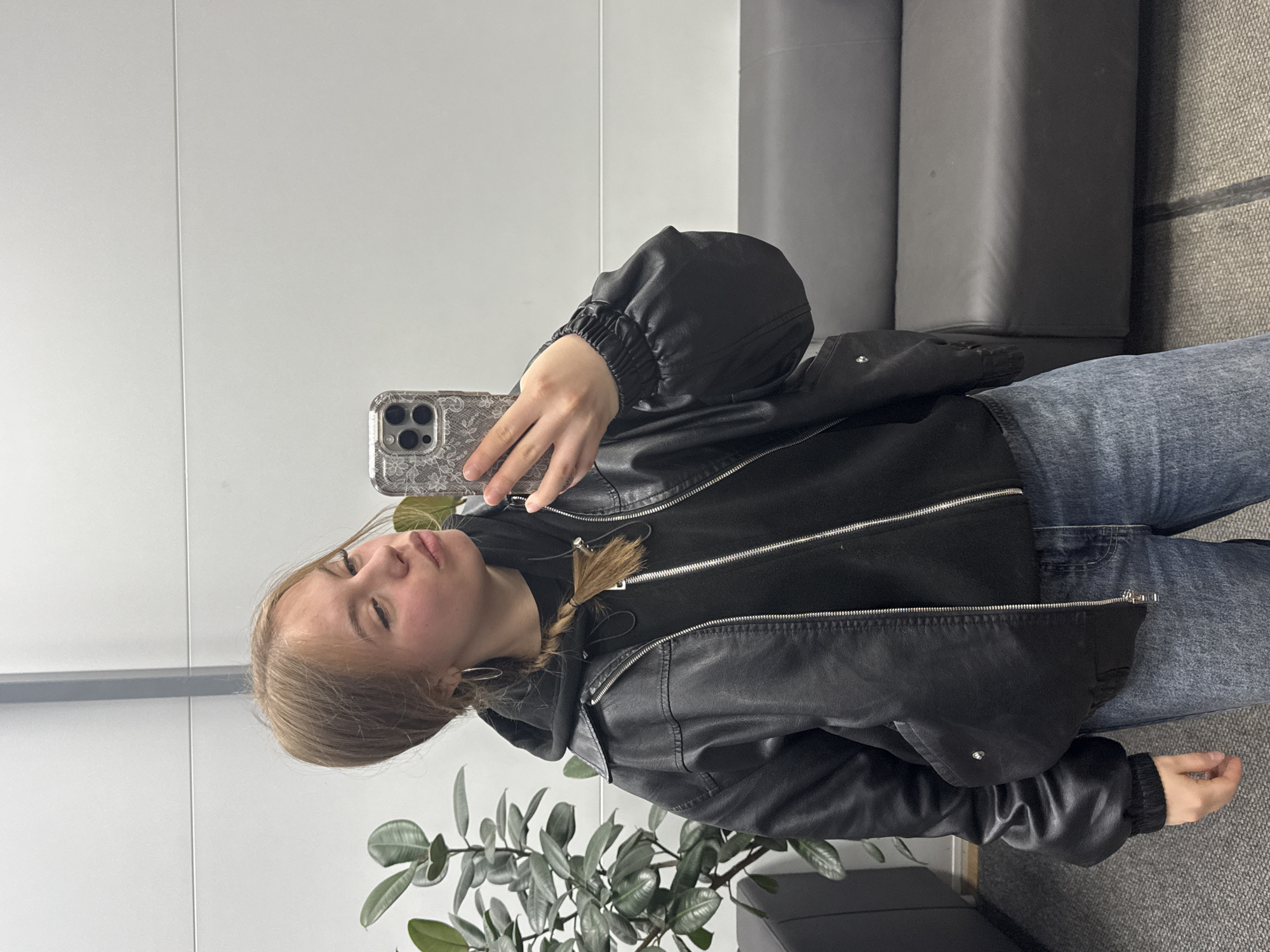 A young woman is captured in a mirror selfie, seen from the chest up, with the image rotated 90 degrees clockwise. She has light brown or blonde hair, styled in a single braid over her left shoulder, and is wearing silver hoop earrings. Her expression is somewhat serious, with slightly pursed lips, as she looks directly towards the phone camera.
She is dressed in a black leather jacket over a black hooded top and blue jeans, visible from the waist down. In her right hand, she holds a smartphone, which has a silver or clear case with a patterned, possibly floral or lace, design. The phone features a multi-lens camera system on its back.
The setting appears to be an indoor space with modern, light grey or white walls. A faint vertical line is visible on the wall, suggesting a panel division or seam. The floor is covered with a dark grey, textured material, possibly carpet. In the background, to the right, a dark grey sofa or bench can be seen. A green potted plant, possibly a rubber plant (Ficus elastica), with large, dark green leaves, is situated in the foreground to the left, adding a natural element to the interior.
The overall scene suggests a casual moment, possibly in a lobby, office, or public waiting area in Chaikovsky, Russia. There is no visible text in the image.