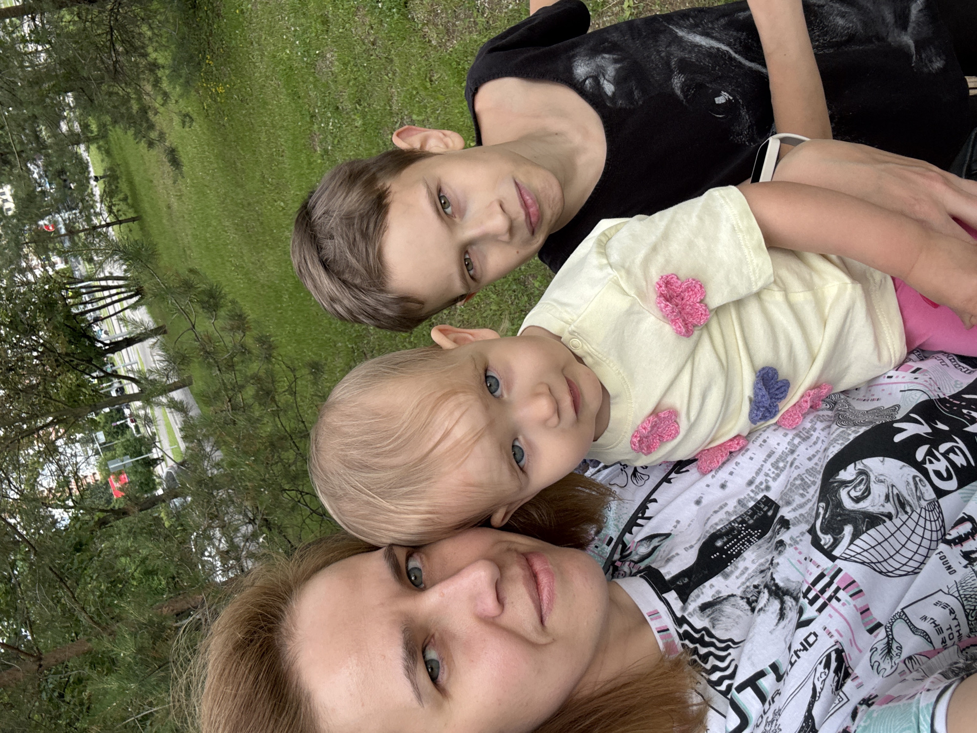 The image is a selfie taken outdoors in Moscow, Russia. In the foreground, three people are visible: a woman, a baby, and a boy. The woman is on the left, holding the baby in her arms. The baby is in the center, wearing a yellow shirt with crochet flowers. The boy is on the right, wearing a black t-shirt with a dog print. They are all looking towards the camera. The background shows a park with trees and a street with cars. The lighting suggests it is daytime.