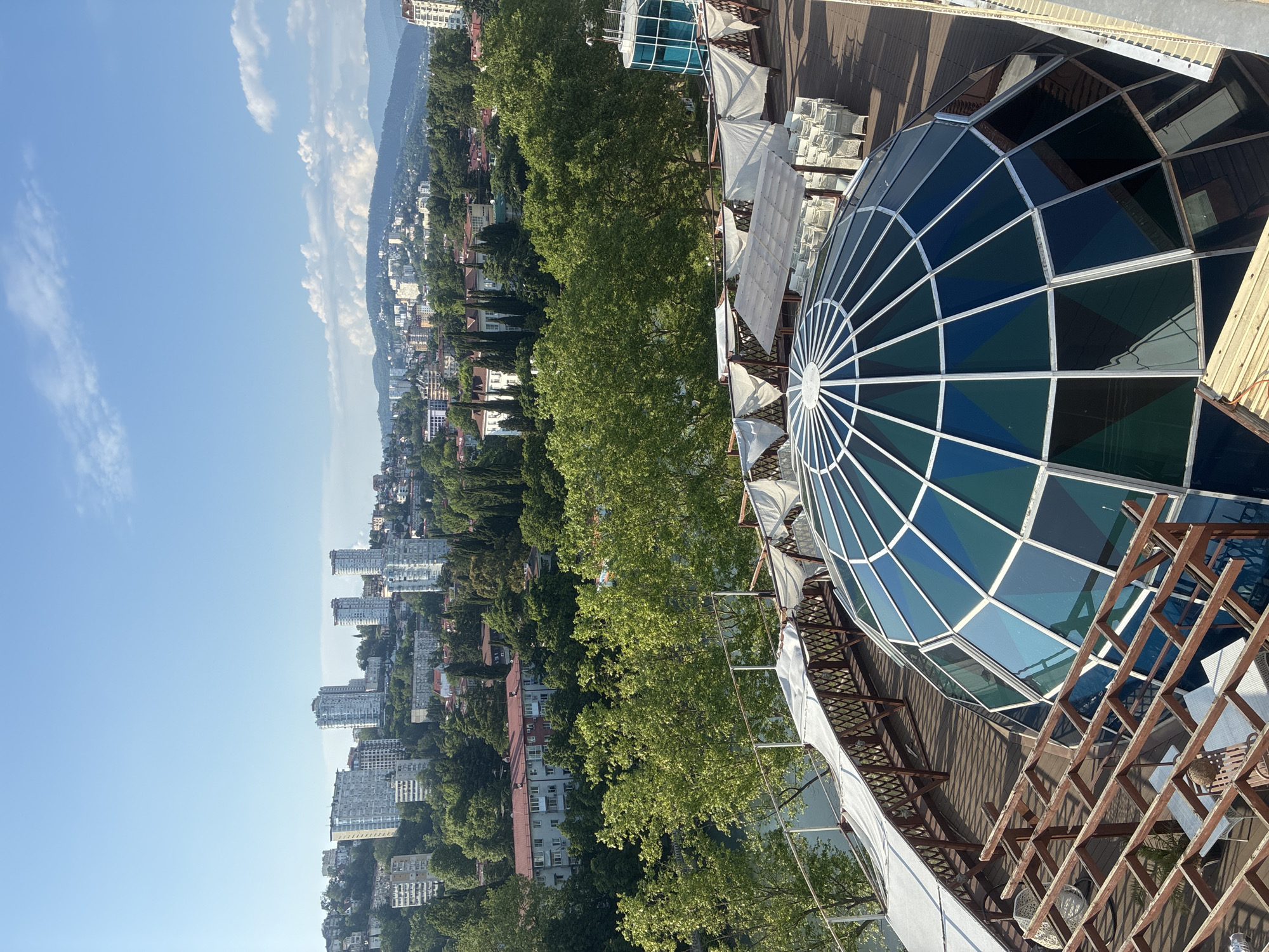 The image displays a rooftop view in Sochi, Russia, featuring a geodesic dome with blue and green glass panels in the foreground. Beyond the dome, a wooden deck area is visible with white chairs and tables under white canopy tents. A water feature, possibly a pool or river, is situated to the left, bordered by a wooden fence and trees. In the background, a cityscape of Sochi unfolds, with numerous tall buildings situated on a hillside dotted with lush green trees. The sky is clear and blue with a few scattered clouds. A blue glass elevator enclosure is also present on the rooftop to the right.
