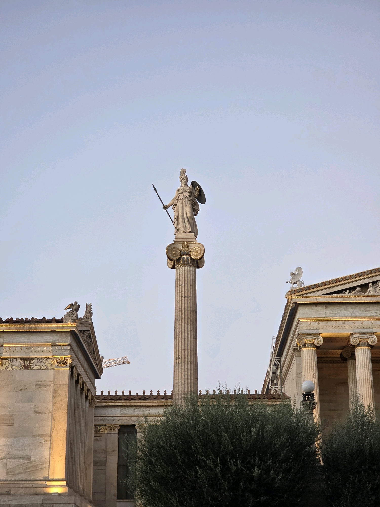 The image depicts the Academy of Athens in Athens, Greece. The central subject is a tall, fluted column topped with a statue of Athena, holding a spear and shield. The statue is made of light-colored stone. To the left and right of the column are classical buildings with marble facades, Doric columns, and ornate friezes. These buildings are illuminated by warm light, suggesting either sunrise or sunset. A dense green bush is visible in the foreground, partially obscuring the lower portions of the buildings. In the background, a light blue sky is visible with a subtle gradient, and a construction crane can be seen in the distance between the buildings. The architectural style is distinctly neoclassical.