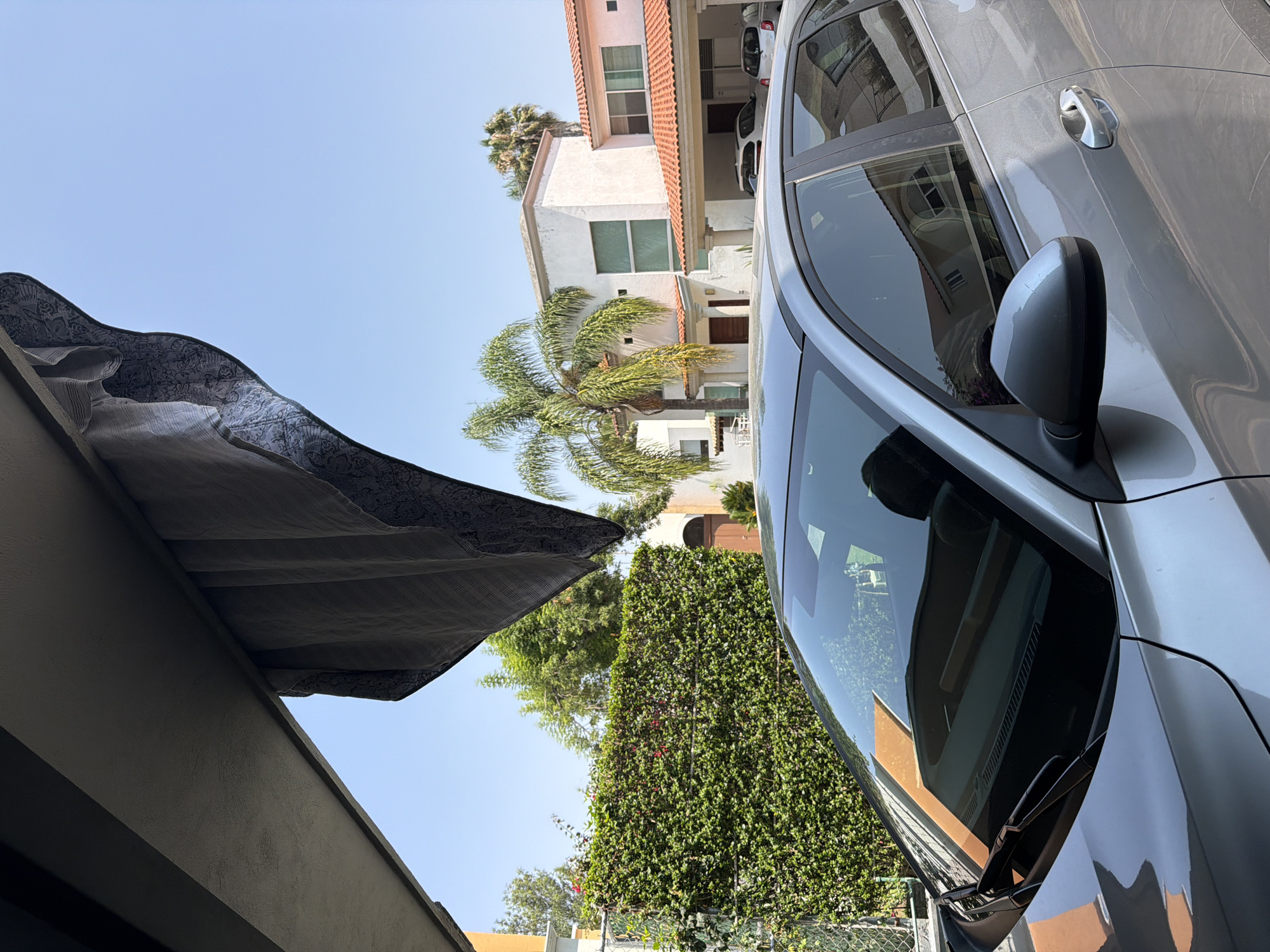 A silver car is parked on a street in a residential area. The car's side mirror is visible. Behind the car, there is a white building with a red tile roof, a palm tree, and a green wall of foliage. The sky is blue. A partially visible awning hangs over the viewer. The location cannot be verified as Puebla, Mexico, from the image alone.