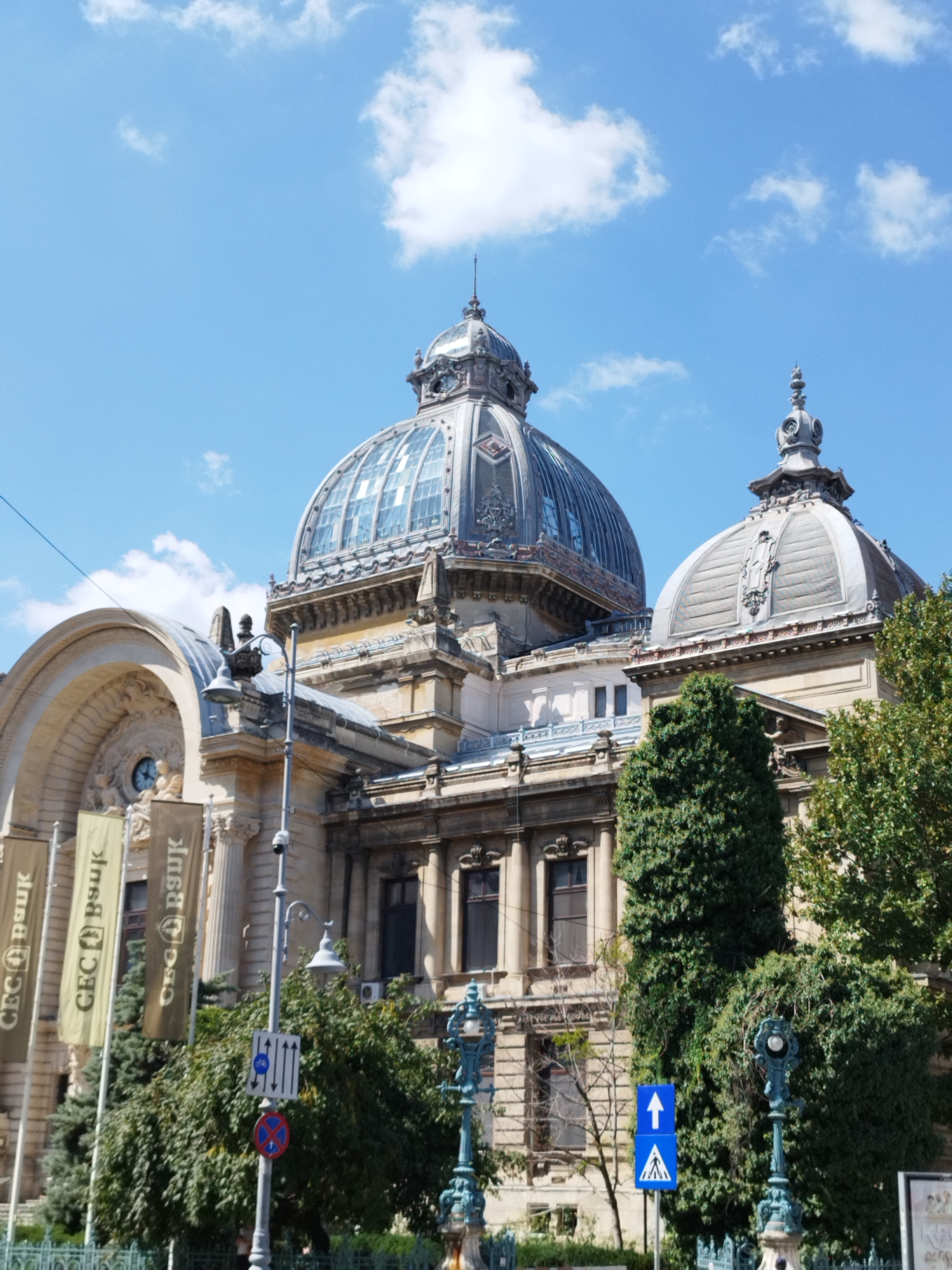 The image shows the CEC Palace in Bucharest, Romania, on a sunny day with a blue sky and scattered clouds. The grand building features prominent domes, classical architecture, and ornate detailing. Several tall, green trees are in the foreground and to the right of the building. To the left of the building, three vertical banners with the text "CEC Bănuk" are displayed. Traffic signs are visible on a pole in front of the trees, including directional arrows and a prohibition sign. The scene is static and appears to be a frontal view of the building's facade. There are no visible people in the image.