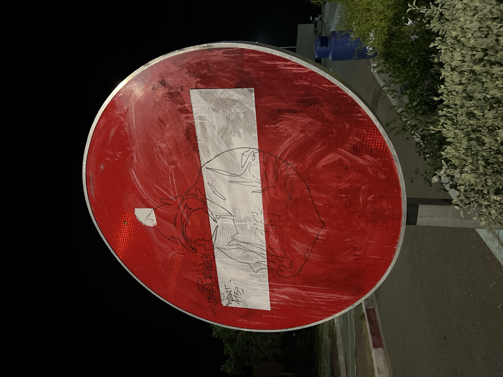 A vandalized "Do Not Enter" sign stands on a street in Jbilate, Morocco. The sign is circular with a red background and a white horizontal bar, partially obscured by white paint and black marker graffiti. The background is dark, suggesting nighttime. A sidewalk and vegetation are visible on the right side of the frame. A blue container sits on the sidewalk.