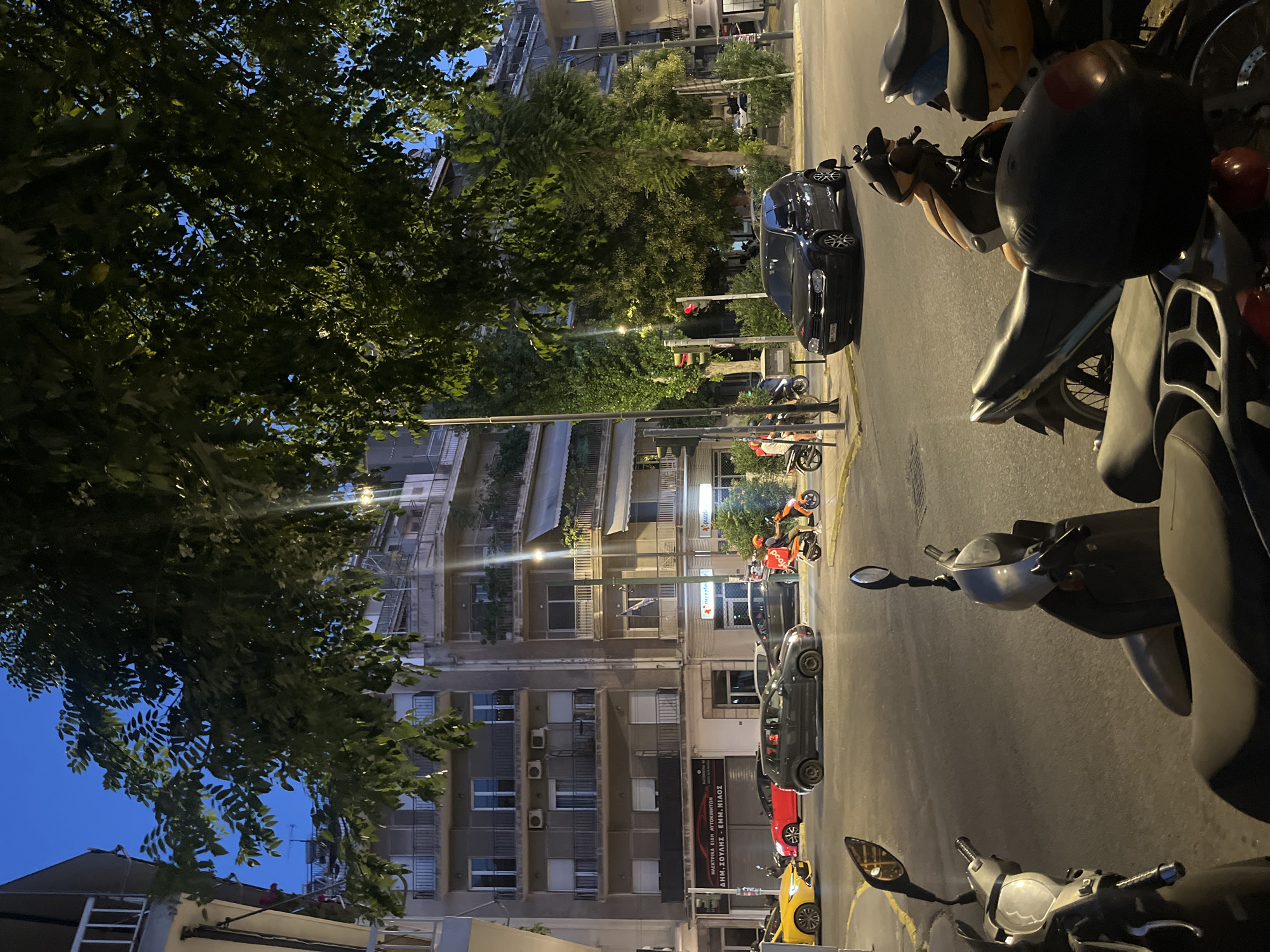 The image depicts a street scene in Athens, Greece, during twilight. In the foreground, several parked scooters are visible. The middle ground shows a street with traffic, including a yellow car, a red car, a dark gray car, and a black Volkswagen Golf. An orange scooter with a food delivery box is also present, ridden by a person in a red helmet. A person on a motorcycle is also visible further down the street. In the background, there are multi-story apartment buildings with balconies. Lush green trees overhang the street, partially obscuring the sky. Traffic lights are visible, with one showing a red signal. The overall atmosphere is that of a bustling urban environment as evening transitions into night.