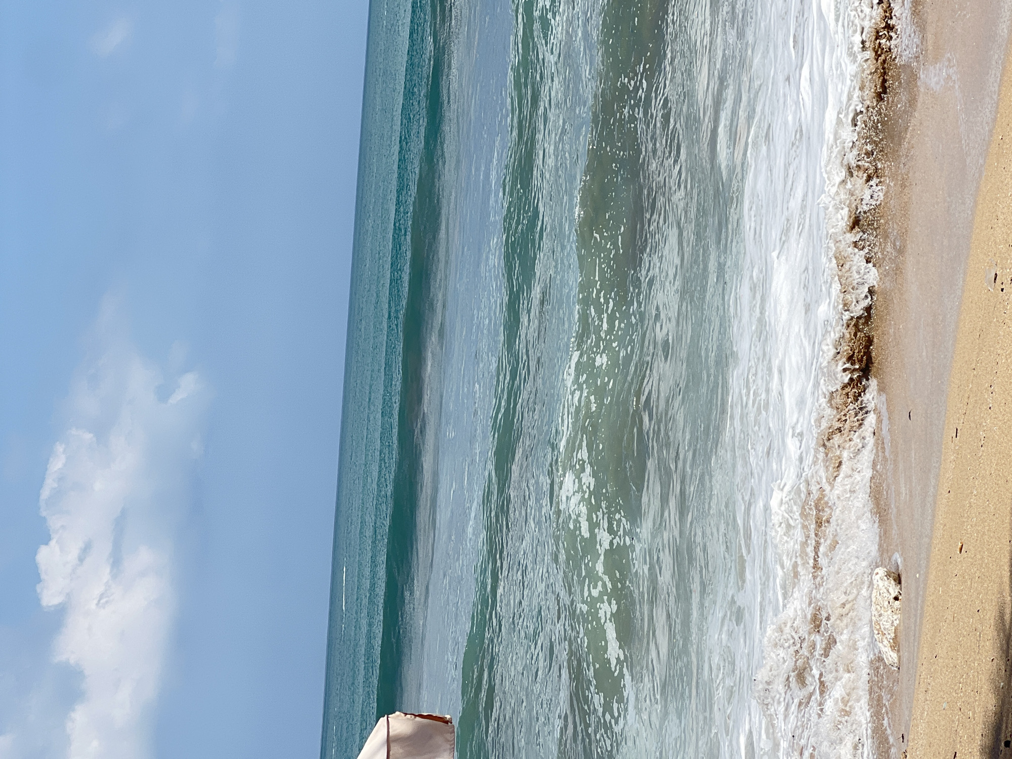 An eye-level, medium shot captures a bright, sunny day at the beach in Kuta, Bali, Indonesia. The ocean dominates the frame, with multiple waves rolling towards the shore. The water transitions from a deep teal in the distance to a lighter, foamy turquoise closer to the beach. On the left side, a portion of a peach-colored beach umbrella is visible, casting a shadow on the sand. The shoreline is made of light brown sand with a few small rocks and debris scattered across it. A single white rock is prominent in the foreground, partially submerged in the breaking waves. The sky above is a clear, pale blue with a few scattered white clouds. The scene conveys a peaceful, tropical beach environment.