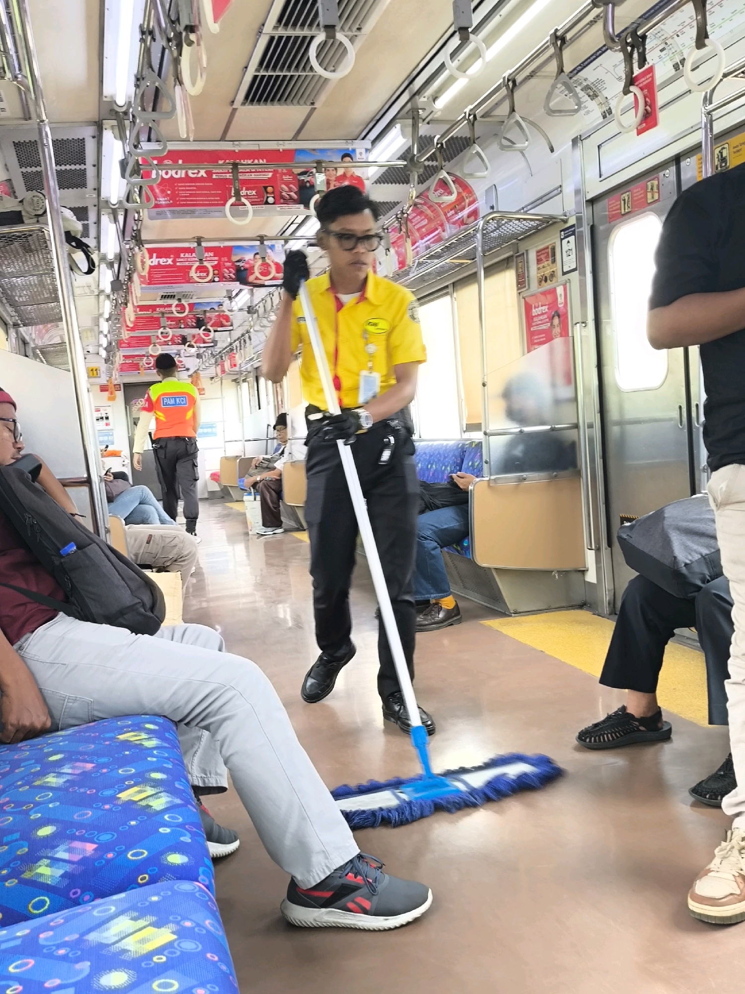 An Indonesian man wearing a yellow uniform and black gloves is mopping the floor of a train carriage. He holds a blue mop with a white handle, and appears to be cleaning the aisle. Other passengers are seated on the blue and patterned seats, some looking ahead and others looking down. The train carriage is illuminated by fluorescent lights. The man in the yellow uniform is focused on his task, while other passengers seem to be going about their journey. The scene depicts a routine cleaning operation within a public transportation setting in Mekarsari, Indonesia.