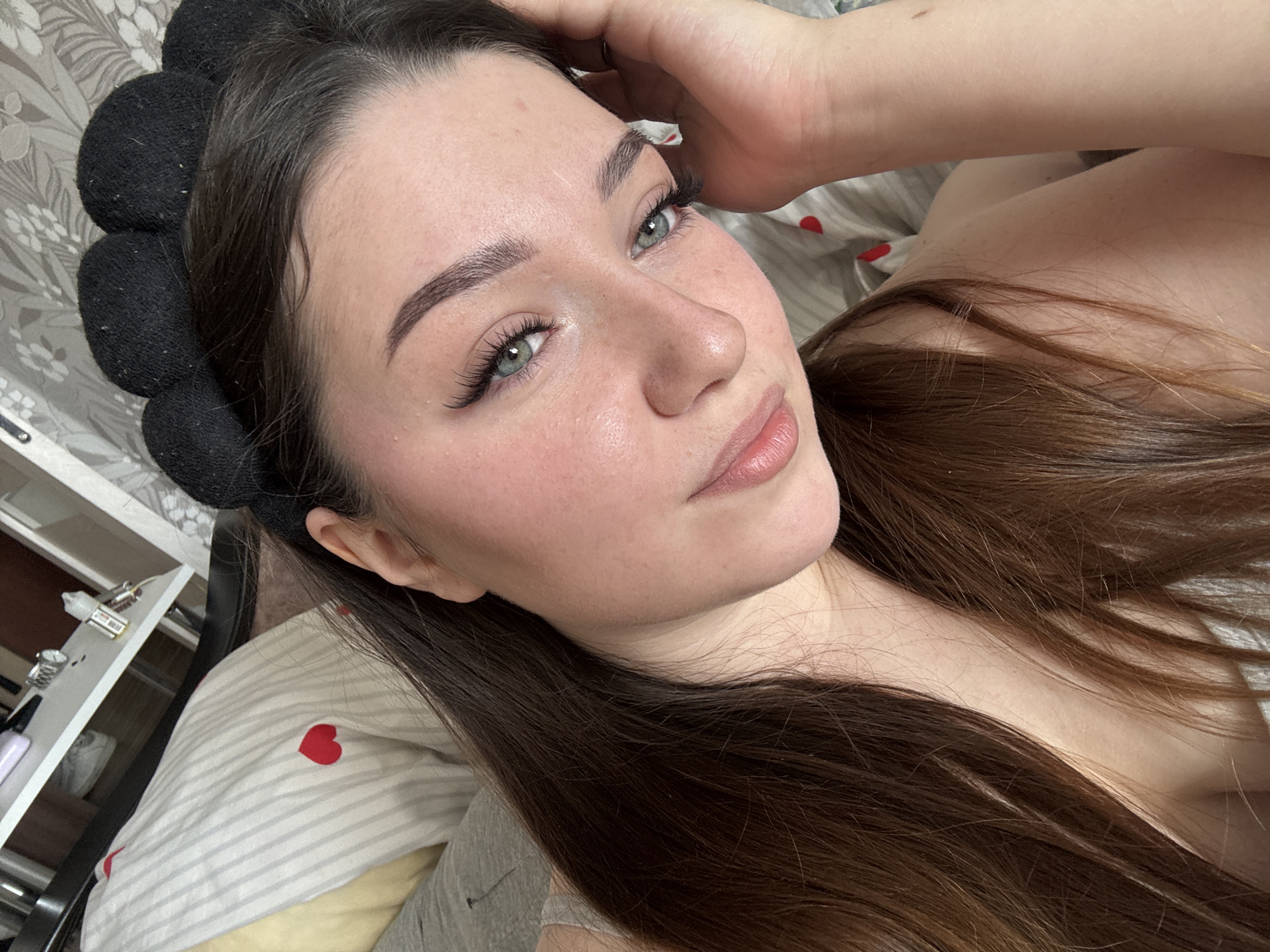 A young woman with long, brown hair and a black mouse-ear headband is lying in bed. She is wearing a grey top. Her face is turned towards the camera, and she is resting her right hand on her cheek. The background shows a bed with striped bedding featuring red hearts, and a white bedside table with items on it. The image is a selfie taken indoors. There is no identifiable information about the location in the city of Perm, Russia.