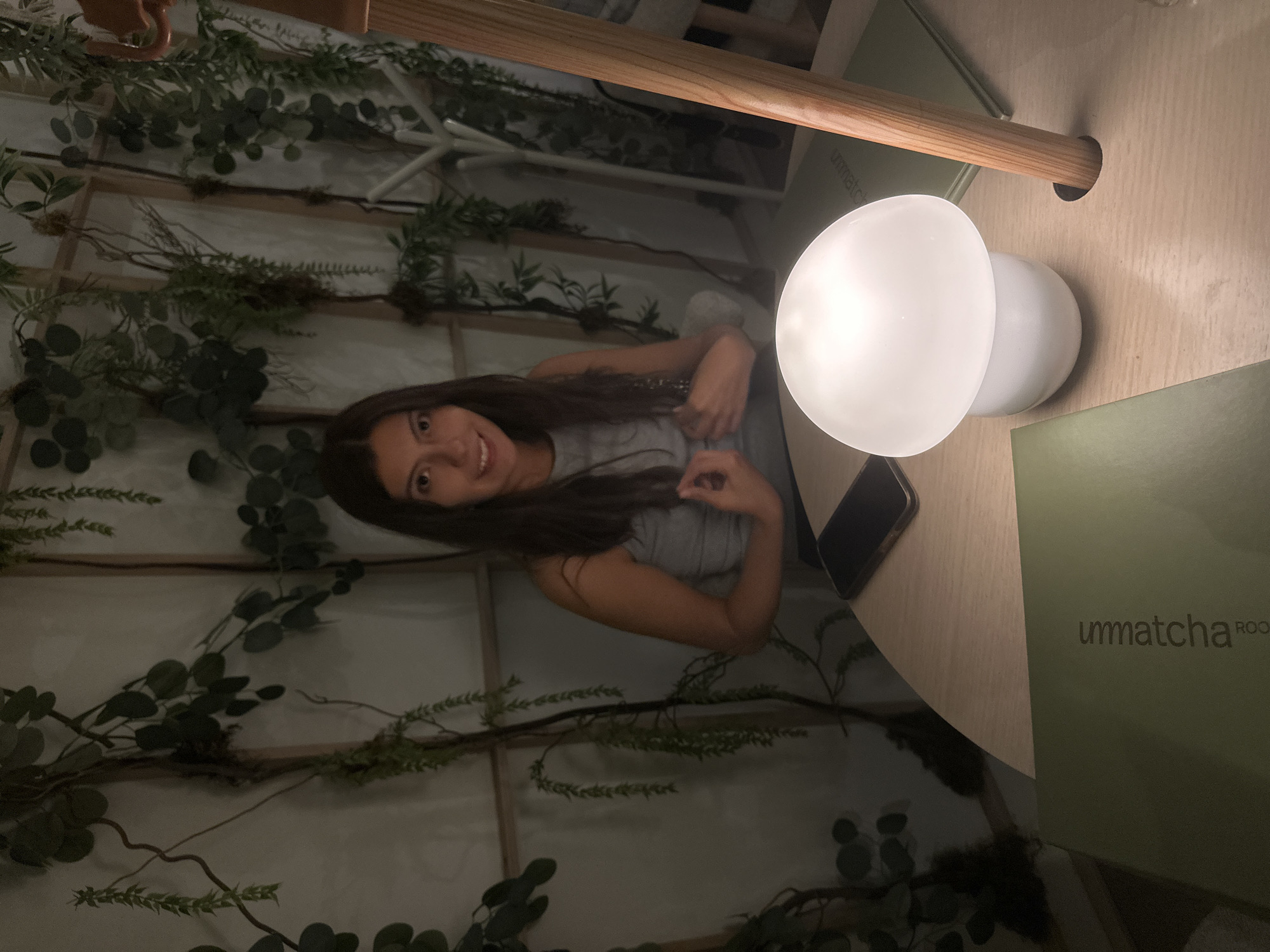 Here's a description of the image:
**Scene:**
The image appears to be taken indoors, possibly at the restaurant "Unmatcha Roo" in the city of Puebla, Mexico. The environment is decorated with artificial foliage creating a garden-like atmosphere.
**People:**
A woman is present in the image. She has long, dark hair and is wearing a light-colored top. She is smiling and appears to be posing for the camera.
**Objects:**
* A lamp with a white globe shade is on the table, providing light.
* A smartphone is lying on the table.
* A menu or book with the word "unmatcha" visible on the cover is on the table.
* A wooden table is in view with a light-colored surface.
* Artificial foliage is used as decoration in the space.
* There is a wooden pole running over the table.
**Actions/Interactions:**
The woman is looking at the camera and smiling. The interaction is a photo being taken.