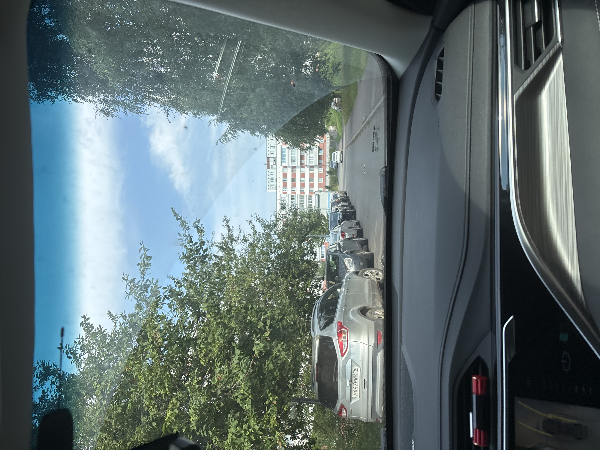 The image captures a view from inside a car, looking through the windshield. The scene depicts a street in Naberezhnye Chelny, Russia, with several cars parked along the side of the road. Mature trees line the street, their green foliage partially obscuring the sky, which is partly cloudy. In the background, a multi-story apartment building with red and white accents is visible. A white car is seen driving away on the road. The foreground shows the interior of the car, including the dashboard and windshield wipers. The overall impression is a snapshot of a residential street on a clear day.