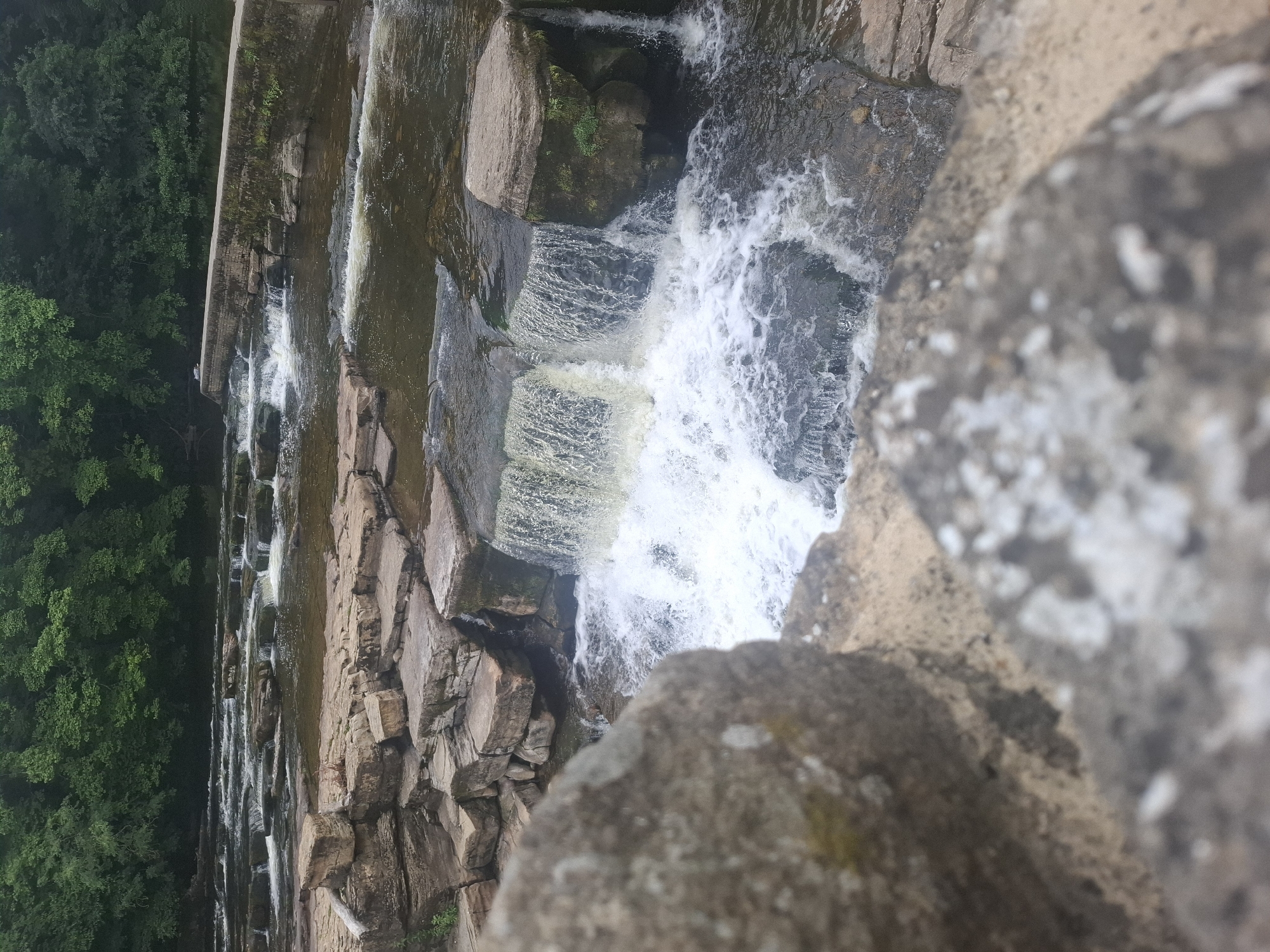 This media file is a photograph of a multi-tiered waterfall cascading over rocks. The scene is dominated by natural elements, with a dense, lush green forest visible on the left side of the frame and large, rough-hewn rocks forming the structure of the waterfall and the foreground. The water is white and frothy as it tumbles down, creating a dynamic contrast with the static rocks and greenery. The image appears to be taken from a high vantage point, looking down at the waterfall. The lighting suggests it might be an overcast day, as there are no harsh shadows, and the overall tone is muted. There is no visible text in the image. The setting is clearly a natural, outdoor environment, likely a park or a nature reserve, given the presence of a constructed waterfall and the well-maintained forest. The focus is on the raw beauty of the water and rocks, creating a sense of tranquility and the power of nature. The composition, with large rocks in the foreground slightly out of focus, adds depth and draws the viewer's eye towards the energetic movement of the water.