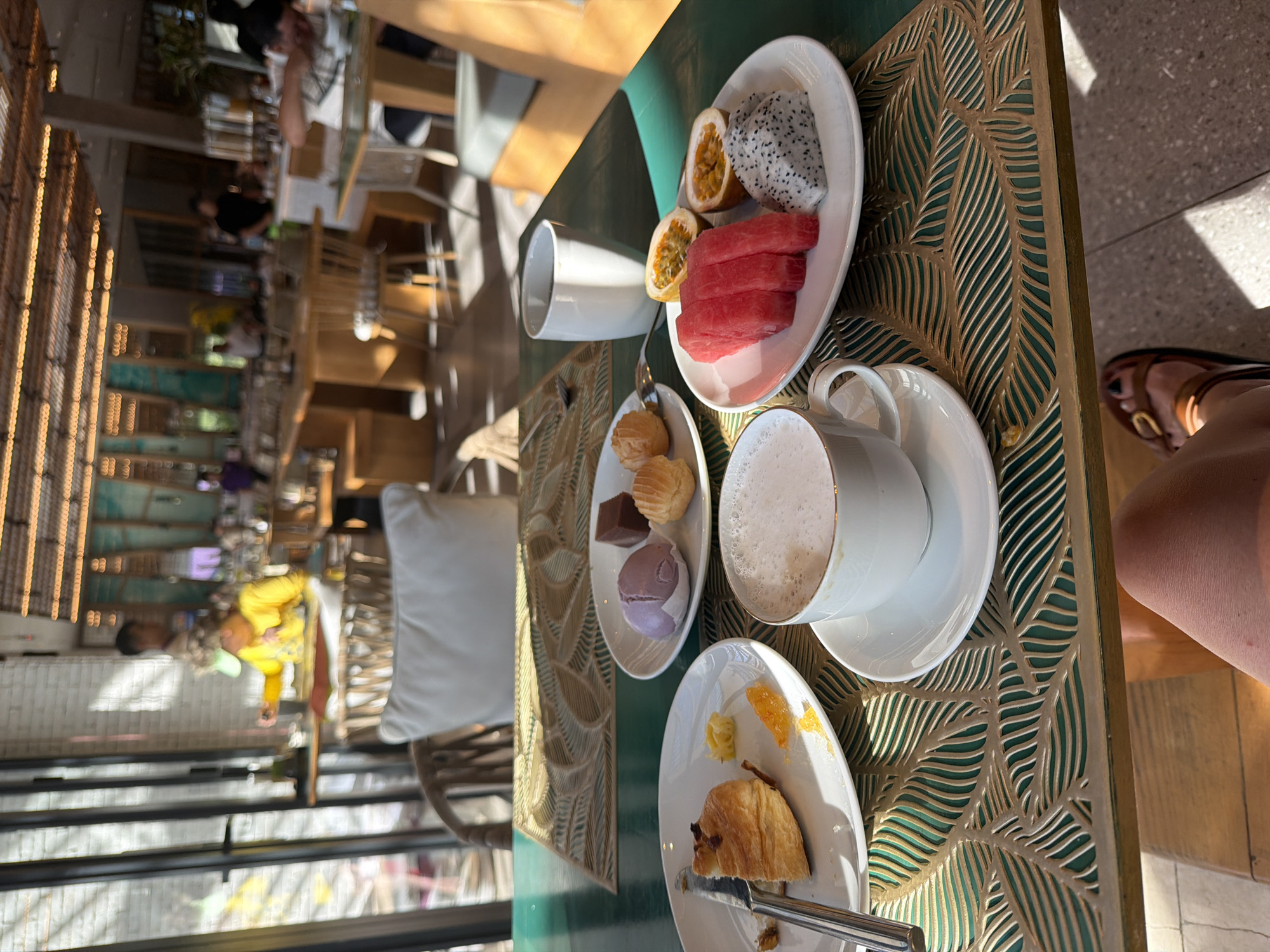 This is a medium shot, eye-level photograph taken indoors at a restaurant in Dong Ninh Hoa Ward, Vietnam, likely during breakfast or brunch. In the foreground, a dark green table displays a meal. On the left, a white plate holds a partially eaten croissant, a knife, and small pieces of orange and yellow fruit. Adjacent to it, another white plate contains a purple mochi, a square piece of dark chocolate, and a small, golden-brown pastry. A white teacup with a frothy beverage sits on a saucer in the center of the table. To the right, a white plate features sliced watermelon, two halves of passion fruit, and a piece of dragon fruit. In the background, blurred figures of diners and restaurant patrons are visible. The restaurant environment appears upscale, with wooden tables and chairs, and decorative elements on the ceiling and walls. Natural light streams in from the left side of the frame, casting shadows across the table and floor.