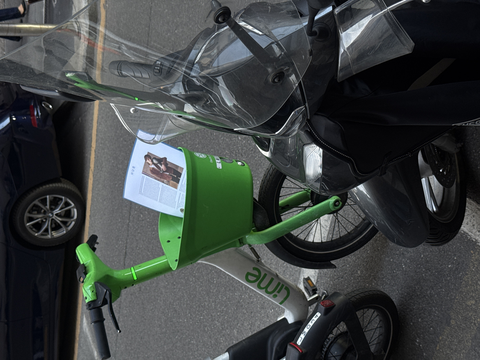 An outdoor, slightly tilted shot captures a street scene in what appears to be an urban environment, possibly Milan, Italy, given the context. The primary subjects are two parked vehicles: a black scooter or motorcycle and a lime green and white electric bicycle or scooter from the company Lime.
The black scooter, positioned towards the top right, features a large, transparent windshield, black handlebars with rubber grips, and a black body. A shiny headlight is visible at the front, along with a dark gray front fender and a front wheel with a disc brake. Part of a black leg cover, possibly with a brand name like "Tucano" faintly visible, is also seen.
To the left and below the black scooter is a bright lime green and white Lime electric bicycle or scooter. It has a green front section with a basket, into which a white piece of paper or flyer with an image of a person (a woman in a stylish jacket) and the word "HUB" is placed. The bike's frame is predominantly white with the "Lime" logo in green. Black handlebars with brake levers are visible, as are foot pedals and its black wheels. The rear fender of the Lime vehicle shows the alphanumeric code "M300A1".
In the background, to the far left, a dark blue car is partially visible, showing its front wheel with multi-spoke silver rims. The ground is asphalt, marked with a faded yellow line, suggesting a road or parking area. The lighting indicates it is daytime. No people are directly present in the scene, and the overall impression is one of parked vehicles awaiting use.
