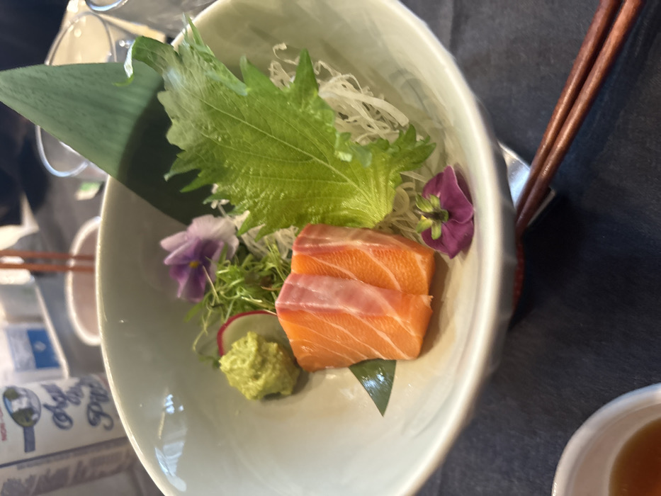 A top-down shot shows a bowl of sashimi. Two slices of salmon rest on a green leaf. A dollop of wasabi, microgreens, radish slices, shredded white vegetables, a large green leaf, and two purple flowers garnish the dish. Chopsticks rest on the bowl's side. Part of a soy sauce container and a small dish of sauce are also visible. The bowl sits on a dark surface.