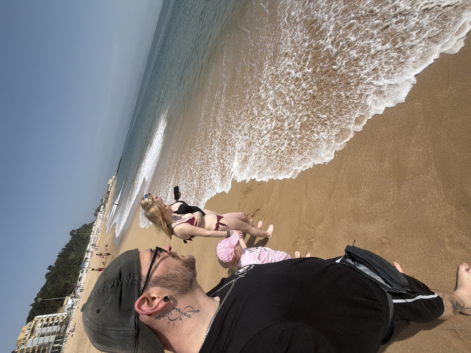 The image shows a sunny beach scene in Oeiras, Portugal. A man wearing a black t-shirt, cap, and glasses is lying on the sand, facing the ocean. He has a tattoo on his neck. Next to him, a child in a pink outfit and hat is also lying on the sand. Further in the frame, a woman in a black and burgundy bikini is lying down, holding a black smartphone. The beach is sandy, with waves reaching the shore. Buildings and trees are visible in the background, indicating a coastal city. People are present on the beach in the distance.