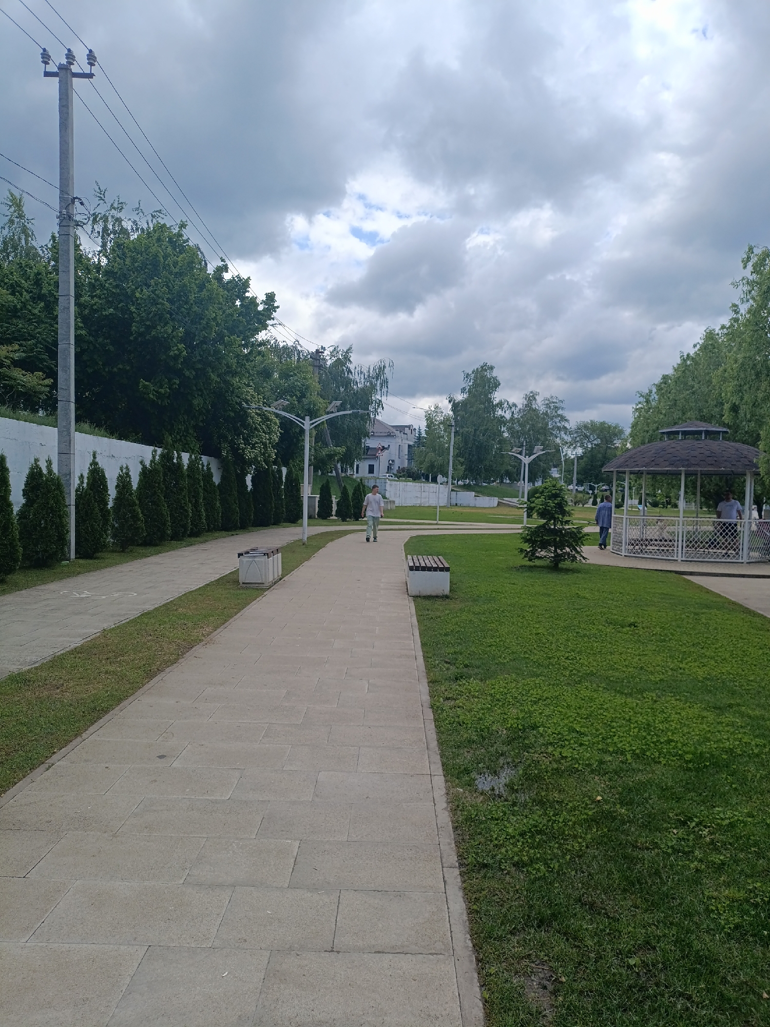 The image shows a paved walkway in a park in Goryachiy Klyuch, Russia. A tall concrete electrical pole stands on the left, adjacent to a white wall lined with rows of coniferous trees. To the right of the walkway is a grassy area with a small evergreen tree, benches, and a gazebo where two people are standing. A man is walking along the paved path away from the viewer. The sky is overcast with grey clouds.