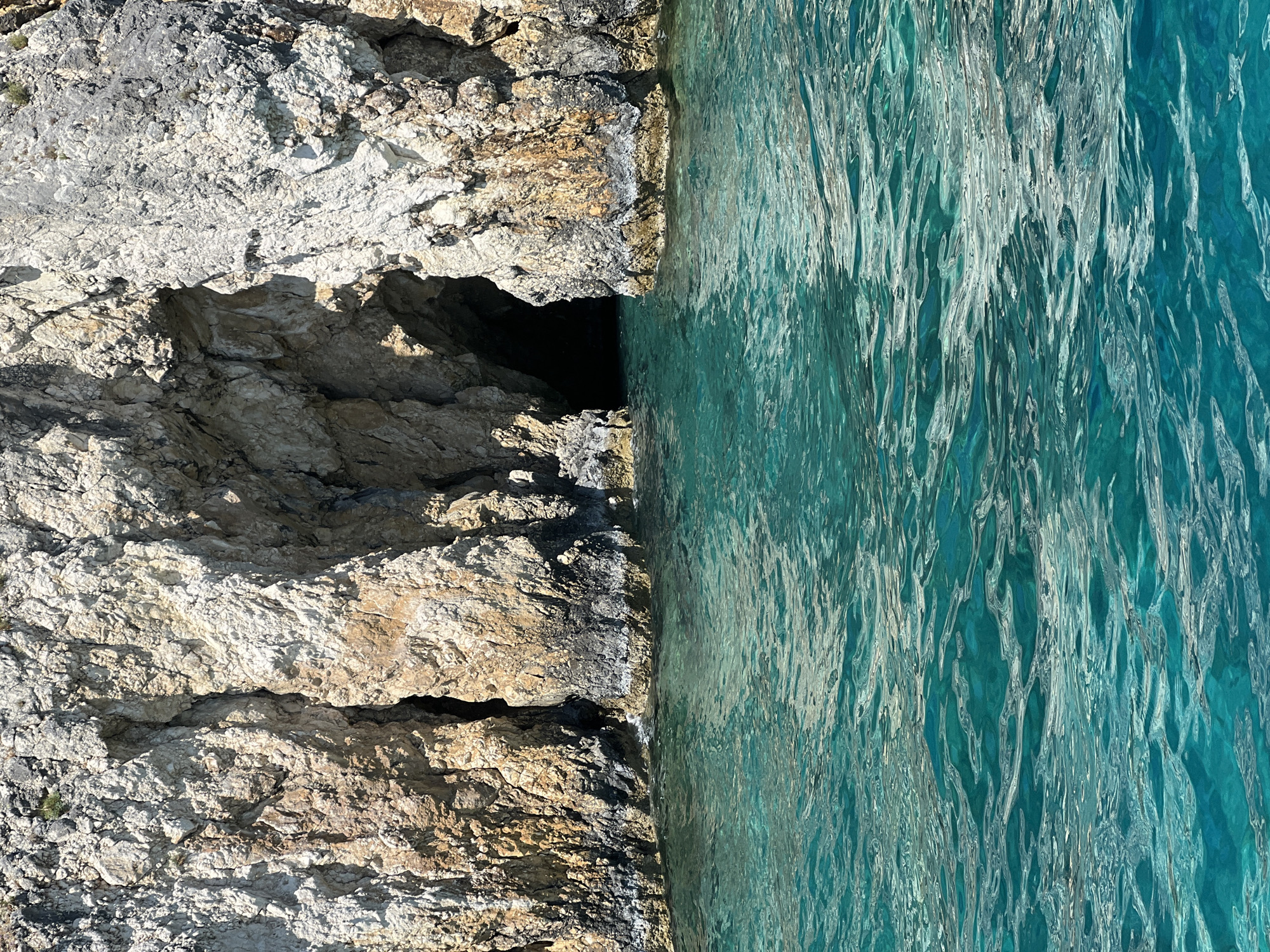 A wide shot captures a natural sea cave carved into a rocky cliff face, with crystal-clear turquoise water lapping at its base. The cave entrance is a dark, shadowy opening, hinting at the depth within. The rocks are layered and textured, showing shades of white, tan, and dark grey, with some patches of green vegetation clinging to the upper ledges. The water in the foreground is calm, with gentle ripples reflecting the sunlight. This scene is located near the city of Laganas Municipal Unit, Greece, likely on the island of Zakynthos, known for its dramatic coastline and sea caves. There are no people or animals visible in the image. The overall impression is one of natural beauty and a serene, untouched environment.