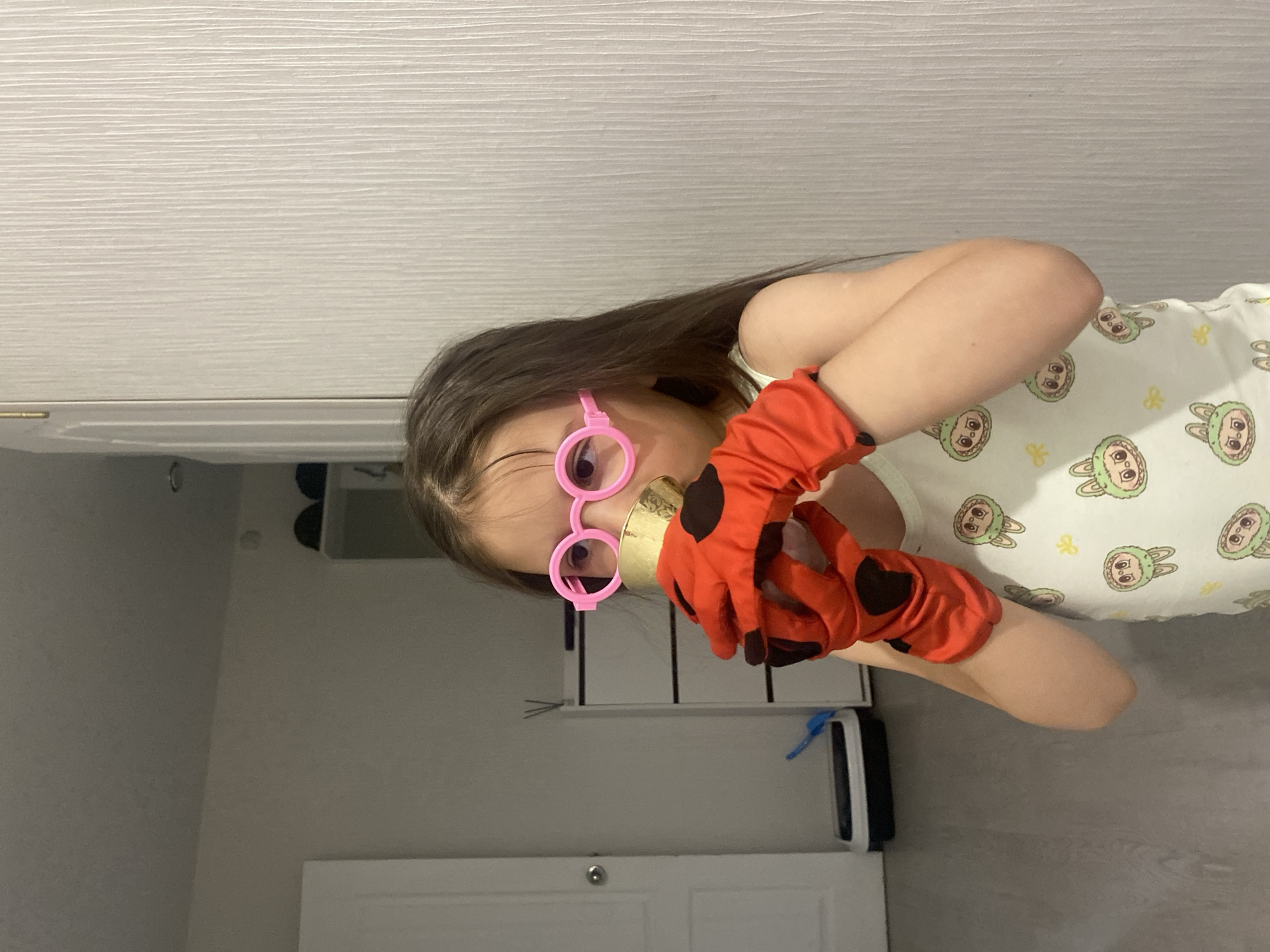 A young girl is captured indoors, dressed in playful attire, looking directly at the viewer. She has long, dark brown hair and wears a light-colored top patterned with cartoon bunny faces and small yellow bow ties. Her hands are covered in red gloves with black polka dots, resembling ladybug gloves, and she holds a golden cylindrical object between them. She is also wearing oversized, pink, circular-framed glasses.
The background suggests a modern home interior in Chaikovsky, Russia. Above her head, a light-toned wall or cabinet with a subtle horizontal wood-grain texture is visible. Below this, a darker greyish-brown wall or surface forms the mid-ground. The lower portion of the image shows a light-colored floor with a wood-like pattern, and a white door or cabinet with a silver knob is partially visible. A blue item rests on a shelf or surface in the mid-right area. The overall scene conveys a moment of imaginative play or dress-up. There is no visible text in the image.