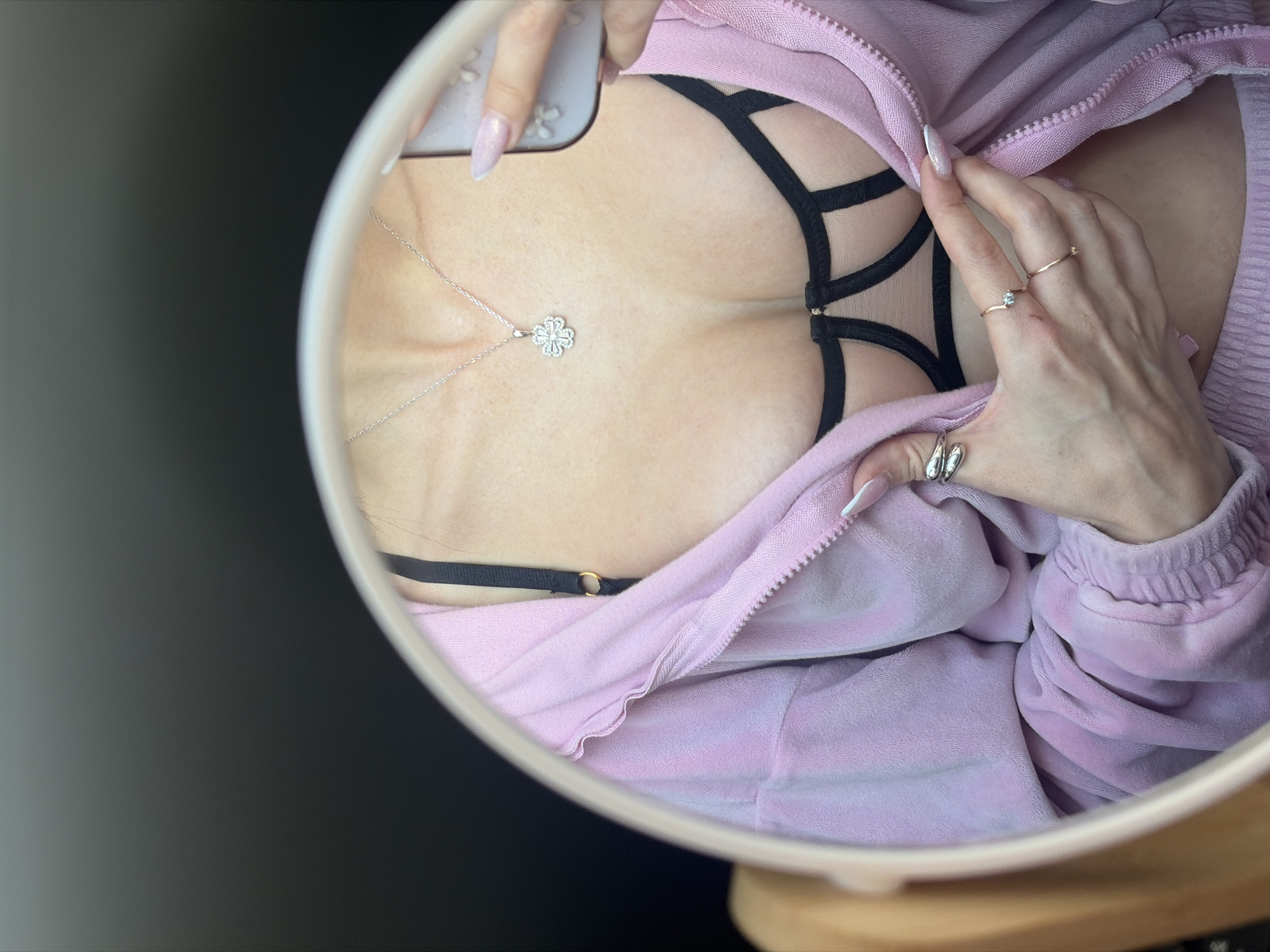 A mirror selfie captures a close-up reflection of a person's upper torso and hands in a round or oval mirror with a light-colored frame. The subject wears a light pink, possibly velour or fleece, zipped hoodie, which is partially unzipped. Beneath the hoodie, a black bra is visible, featuring mesh panels and multiple straps that form geometric patterns across the chest. A delicate silver chain necklace with a sparkling, floral or clover-shaped pendant rests on the chest.
The left hand holds a smartphone, partially visible in the reflection, and has long, light-pink, glittery manicured nails. The right hand, adorned with French-tipped manicured nails, rests on the hoodie near the chest. It displays a plain gold ring, a gold ring with a small solitaire diamond, and two silver rings on the thumb. The background reflected outside the mirror's frame is dark and undifferentiated, suggesting an indoor setting, possibly with low lighting or dark furnishings. No visible text is present in the image. This mirror selfie is contextualized as being taken in Saint Petersburg, Russia.
