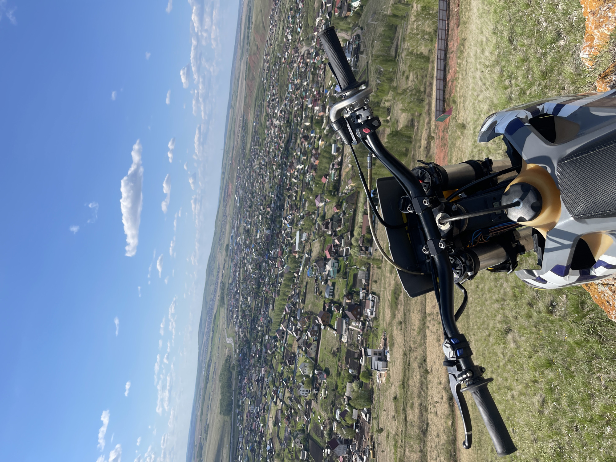 View from the handlebars of a motorcycle parked on a grassy hill overlooking a residential area. The residential area contains numerous small buildings with varying roof colors, interspersed with green trees and vegetation. Beyond the residential area is a vast expanse of flat land with patches of agriculture and distant structures. The sky is blue with scattered white clouds. The motorcycle is partially visible, including the handlebars, controls, and part of the fuel tank and front fairing. Дрокино, Russia.