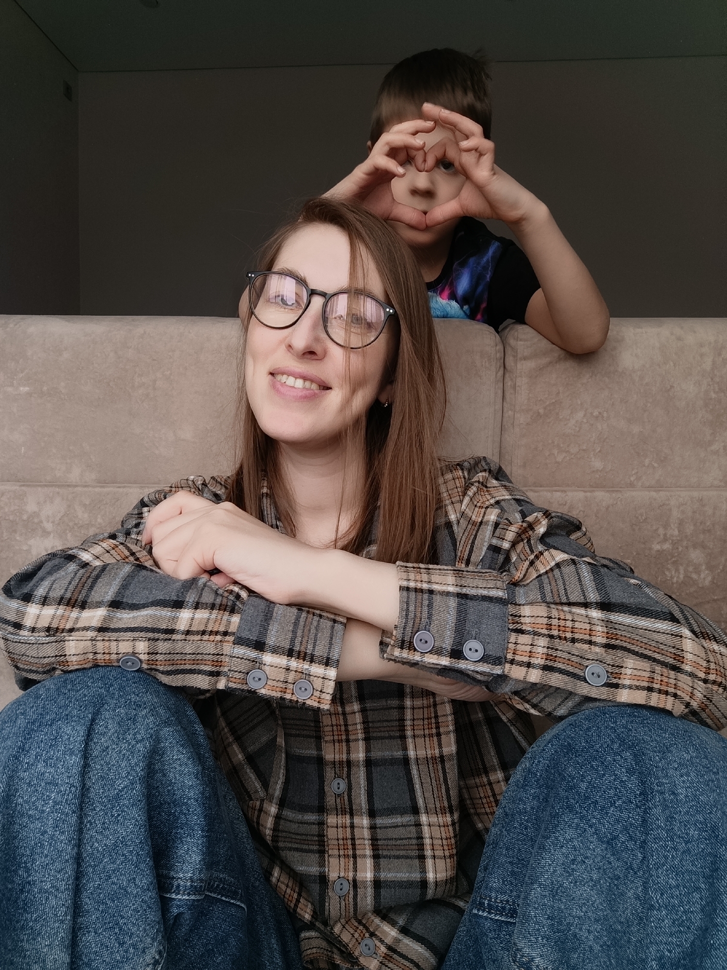 A woman is seated cross-legged on a beige couch, wearing glasses, a plaid shirt, and blue jeans. She is smiling and has her arms crossed, resting on her legs. Behind her, a child is making a heart shape with his hands, framing his face. The background is a plain, dark gray wall. The location is Chaikovsky, Russia.