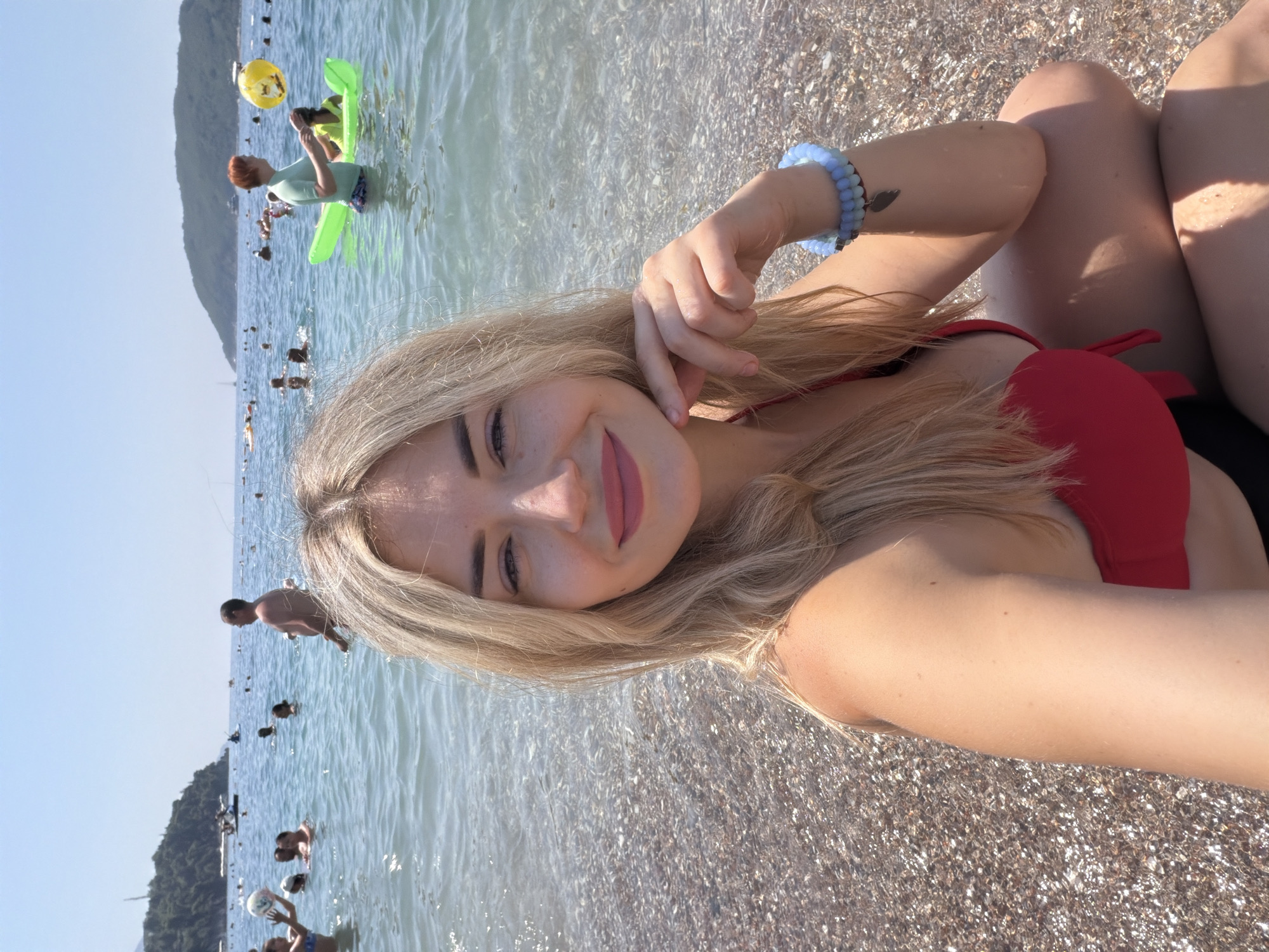 The image shows a woman with blonde hair, wearing a red bikini, sitting on a pebble beach. She is looking towards the camera with a smile. In the background, there are many people swimming and playing in the clear blue water of the Adriatic Sea. There are also some boats and a hilly coastline visible. This appears to be a scene from a beach in Budva, Montenegro, a popular tourist destination known for its beautiful beaches and vibrant atmosphere. The water is calm and inviting, and the sky is clear and sunny, suggesting a pleasant day for outdoor activities.