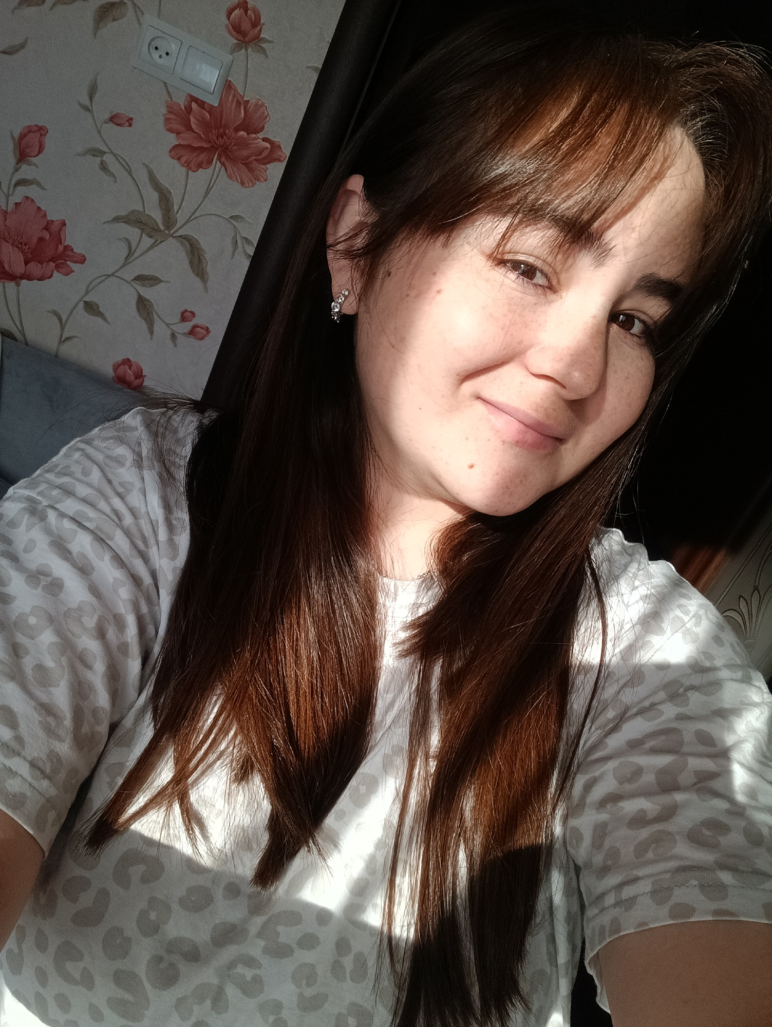 A selfie of a young woman with brown hair and freckles, wearing a light-colored patterned T-shirt. She is smiling slightly. An earring is visible in her left ear. Behind her is wallpaper with a floral design, and an electrical outlet is visible on the wall. The right side of the image is darker, suggesting the presence of a wall or doorway. The sunlight casts shadows on her face and shirt. There is no evidence to suggest this image was taken in the city of Noviy, Russia.