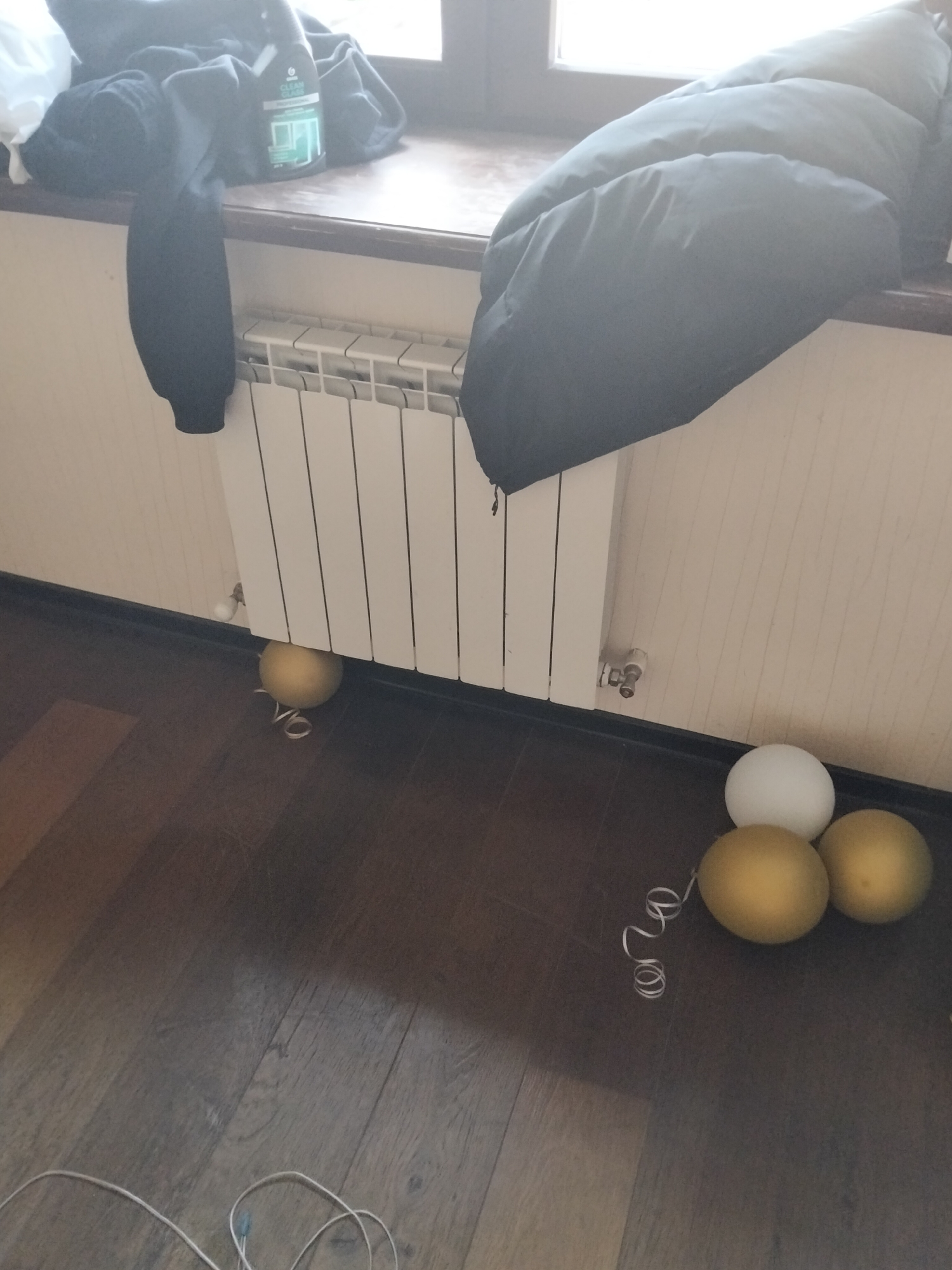 The image shows a radiator in front of a windowsill, with some clothing items draped over the radiator and windowsill. On the floor in front of the radiator are three balloons, two gold and one white, with a white ribbon curled around one of the gold balloons. There is also a white wire lying on the floor. The scene appears to be indoors, in a room with wooden flooring. The location is Unknown, Unknown.