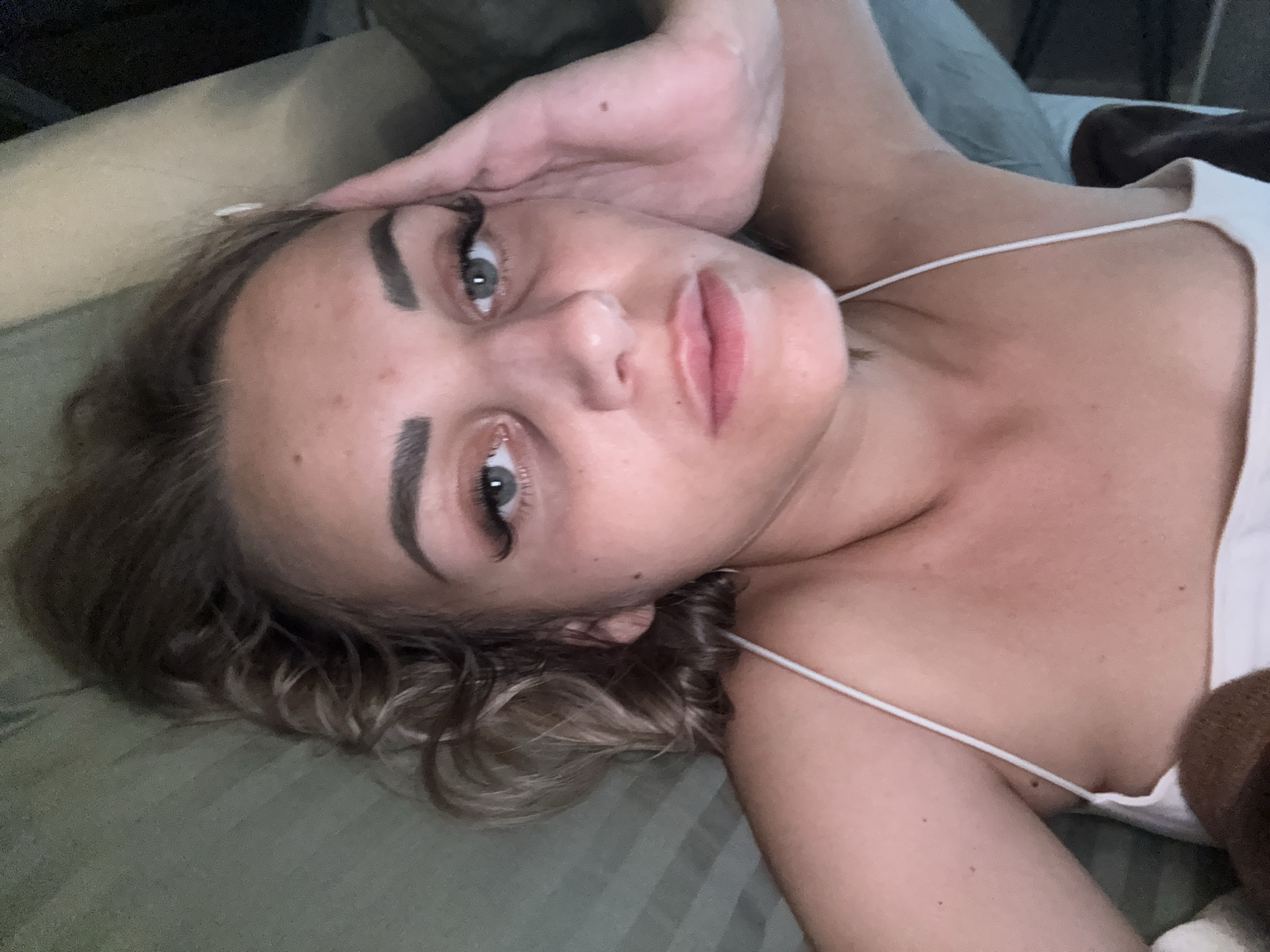 A young woman with blonde, curly hair and prominent eyelashes is lying on her back, looking upwards with a relaxed expression. She is wearing a white spaghetti strap top. Her right hand is raised and resting on her forehead. The background appears to be a bed with green and grey striped bedding. There is no indication of the image being in Yuzhno-Sakhalinsk, Russia.