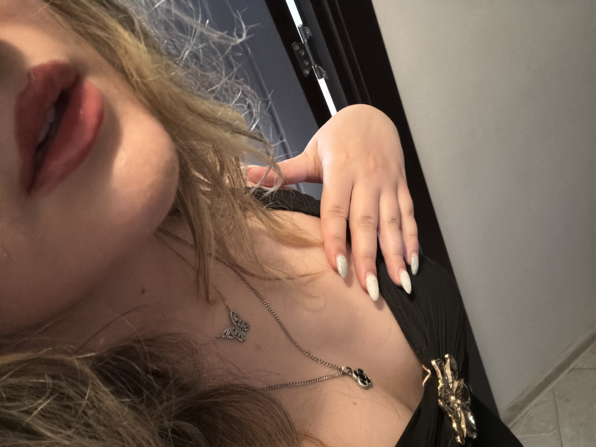 The image is a close-up shot of a person's décolletage, focusing on their chest, neck, and mouth. The person is wearing a black dress with a decorative gold embellishment on the bust. Two silver necklaces are visible: one with a butterfly pendant and another with a black pendant. The person has manicured nails, some painted with glitter and others in a solid white color. Their hair is blonde and appears somewhat disheveled. The background is out of focus, but appears to be an indoor setting with dark horizontal elements and a bright light source. There is no identifiable information within the image to suggest a specific location in Plovdiv, Bulgaria.