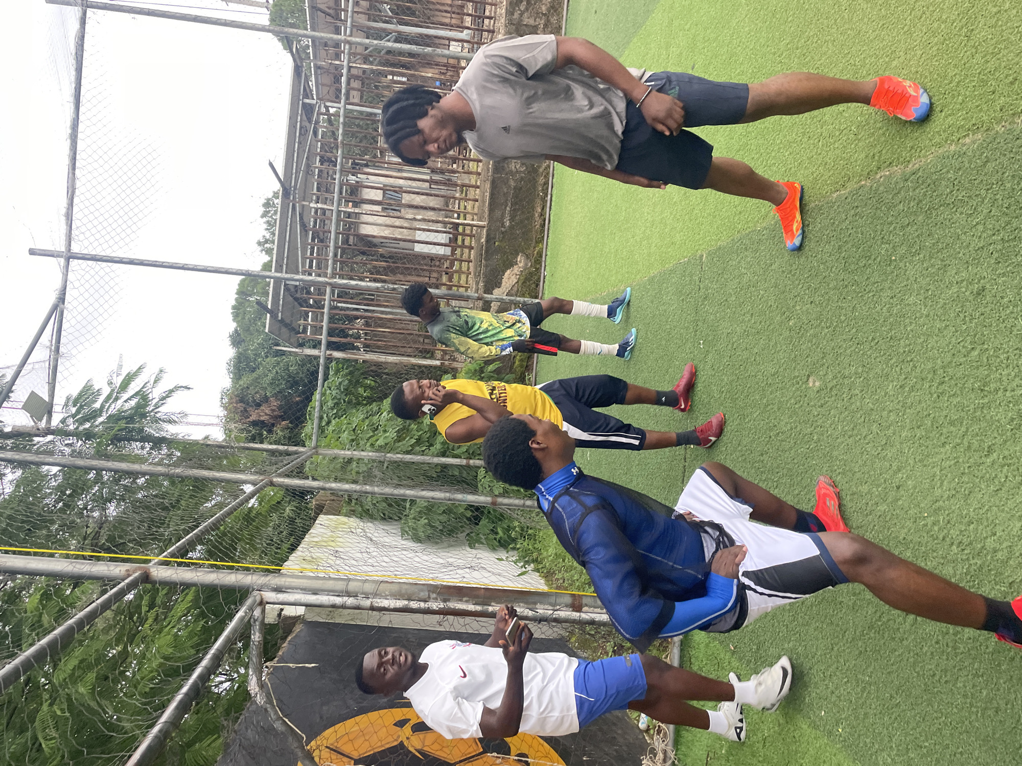 A group of five young men are gathered on an artificial turf field, likely a sports facility, in Jos, Nigeria. The field is enclosed by a chain-link fence, with some trees and buildings visible in the background. The sky is overcast.
The man on the far left wears a white polo shirt with a maroon logo and blue athletic shorts. He is holding a smartphone in his left hand and appears to be speaking.
In the center, a man in a yellow t-shirt and black shorts is holding a smartphone to his ear and talking.
To his right, another man, seen from the back, wears a blue long-sleeved athletic shirt and white shorts with black and white trim. He is in a crouched position, leaning forward with his right hand on his thigh.
Further to the right, a man in a green and yellow tie-dye long-sleeved shirt and black shorts stands with his head bowed. He is wearing blue soccer cleats and white socks.
On the far right, a man with dreadlocks wears a grey t-shirt and dark athletic shorts. He is looking downwards and has a bracelet on his left wrist. He wears bright orange and blue soccer cleats.
The overall scene depicts a casual gathering, possibly before or after a training session or game. The presence of athletic wear and the artificial turf suggest a sports context.