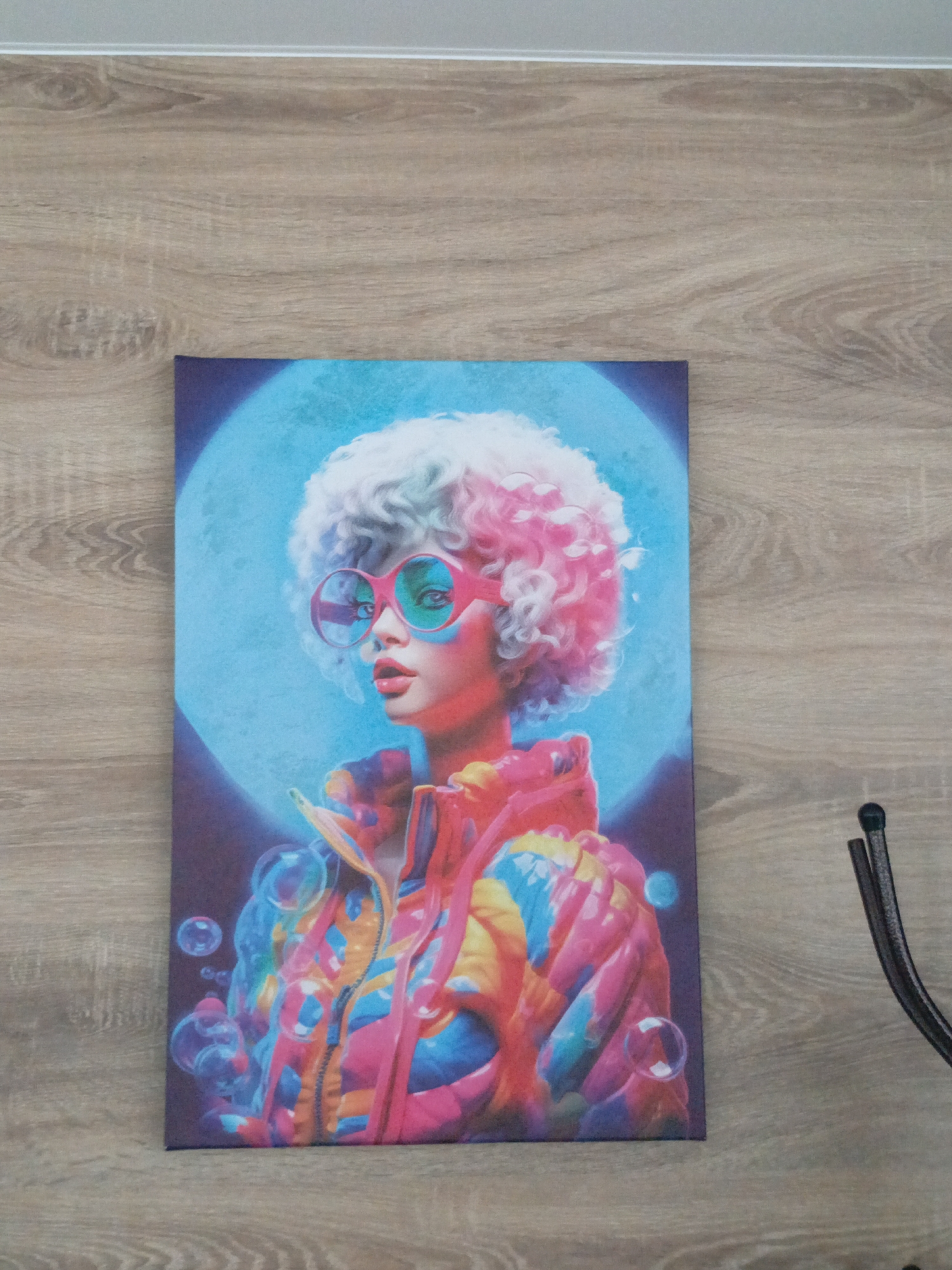 A colorful canvas print hangs on a light brown, wood-grained wall. The print features a stylized portrait of a woman with a white, curly afro tinged with pink, wearing large, pink-rimmed glasses with light green lenses. She is depicted against a blue background with a darker, almost shadowy lower portion. The woman wears a brightly colored jacket with hues of orange, yellow, pink, and blue. Bubbles are interspersed around the bottom part of the image. To the right of the canvas is a portion of a black metal object, possibly a chair or other piece of furniture. The location and city are not discernible from the image.