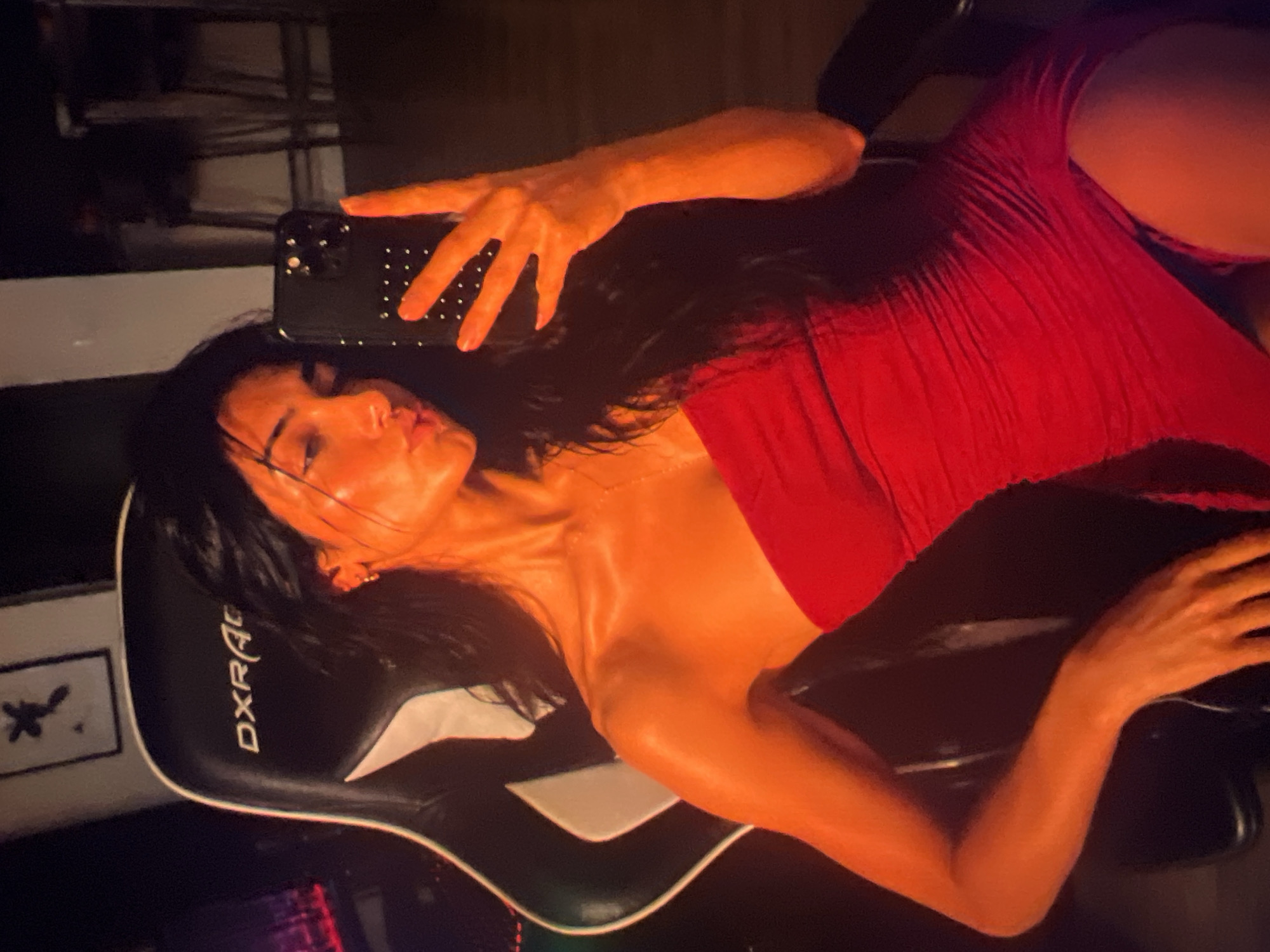 A young woman with dark hair is taking a selfie in a red strapless dress. She is sitting in a black and white gaming chair. The background is dark and appears to be a room with furniture.