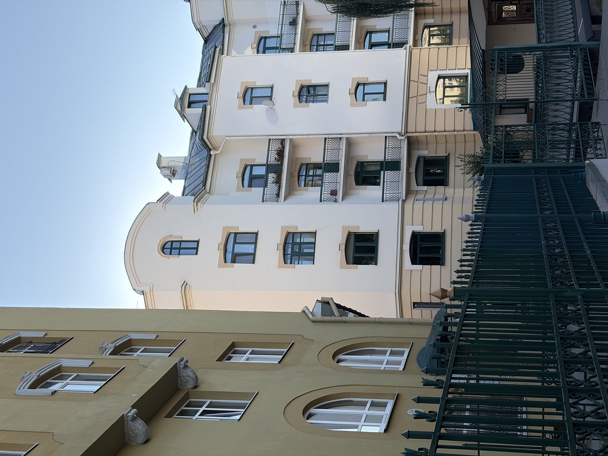 The image captures a daytime exterior view of a residential building in Minsk, Belarus. The building features a light-colored facade with numerous windows, many of which have dark green frames. Several balconies with black metal railings are visible. To the left, another building with a yellowish facade and arched windows is partially visible. A dark green metal fence runs across the foreground, separating the viewer from the buildings. The sky is clear and blue.