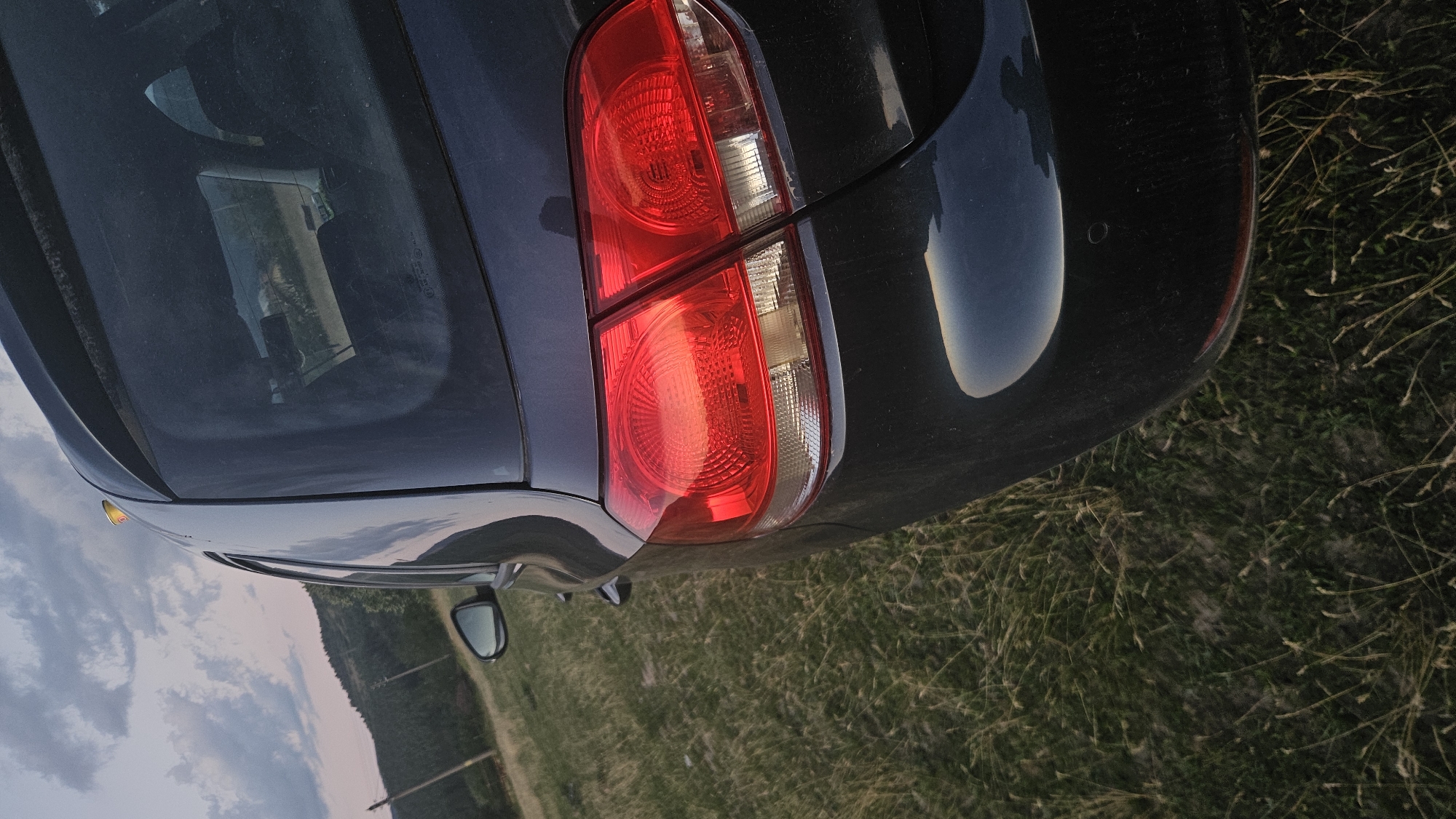 A close-up, rear-quarter view of a dark-colored hatchback car is shown, parked outdoors on a grassy area. The setting appears to be rural, with a hillside covered in trees and vegetation visible in the background, along with a cloudy sky at dusk or dawn. The car's taillights are illuminated, casting a red glow. The rear bumper and a portion of the rear window are also visible. The ground is covered in dry grass and some scattered weeds. There is a small object, possibly a can, visible on the roof of the car, near the rear spoiler. No people are present in the frame.