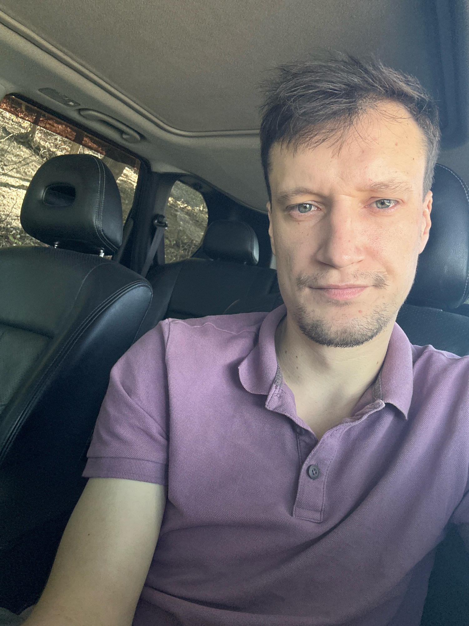 This is a medium shot, taken from inside a car, of a man in his late 20s to early 30s. He has light skin, short brown hair, and a light stubble beard. He is wearing a purple polo shirt. The man is looking directly at the camera with a neutral expression. The background shows the interior of a car, including black leather seats, part of the sunroof, and the headrests of the seats behind him. Through the windows, trees and foliage are visible, suggesting an outdoor setting, possibly a wooded area. The sunlight filtering through the trees and car windows indicates it is daytime. The location is Makiv, Ukraine. No visible text is present.