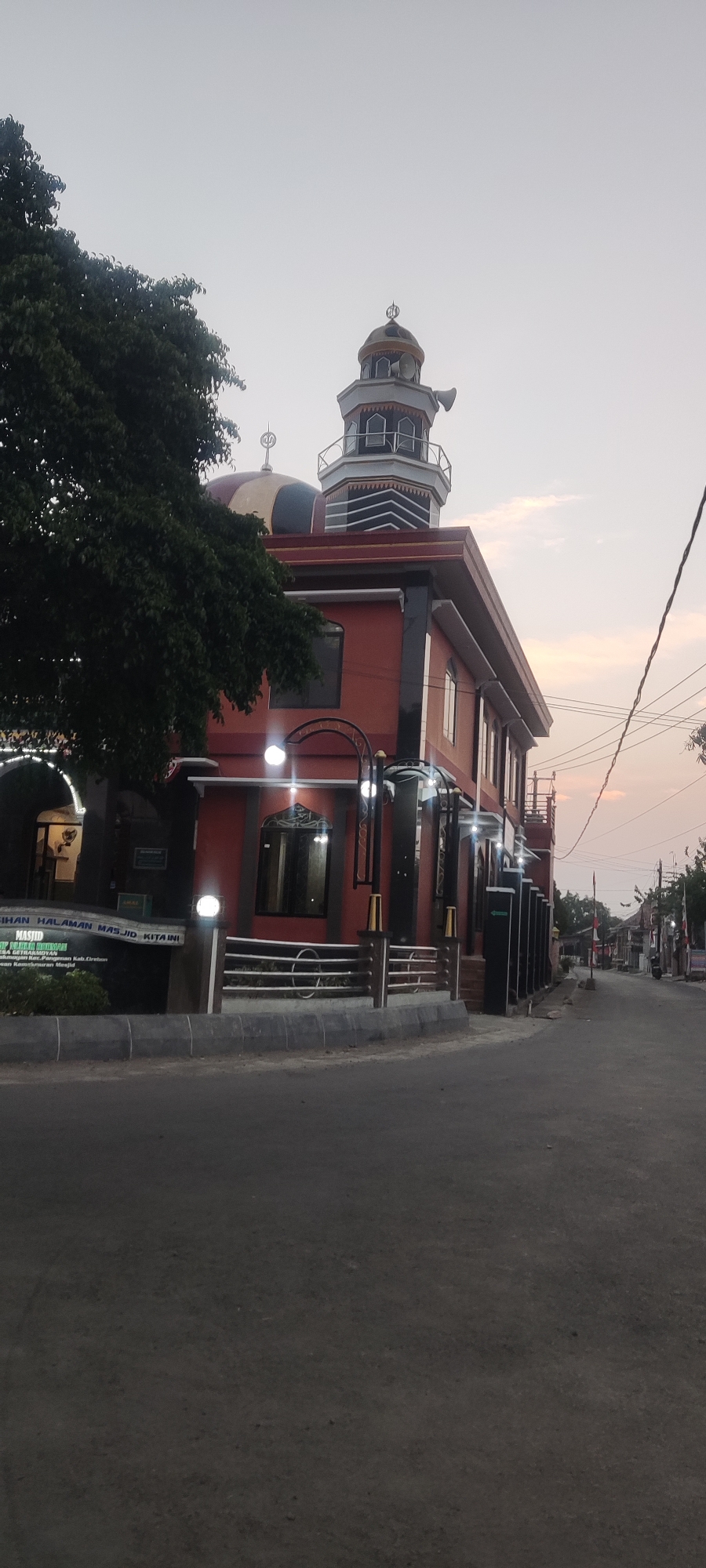 The image depicts a mosque in Getrakmoyan, Indonesia, captured from a low-angle perspective on an overcast day, possibly during dawn or dusk. The mosque, painted in a vibrant orange hue, features a distinct dome and a tall minaret with loudspeakers. A large tree with dark green foliage is situated to the left of the building. The mosque is fronted by a low fence and a paved pathway. Beyond the mosque, a street lined with houses and flags can be seen, with a motorcycle parked on the right side of the road. The sky is a pale orange and grey, indicating the time of day. A signpost at the entrance of the mosque reads "MASJID NURULLAH DRESKMURBAN" and "DESA GETRAKMURBAN."
