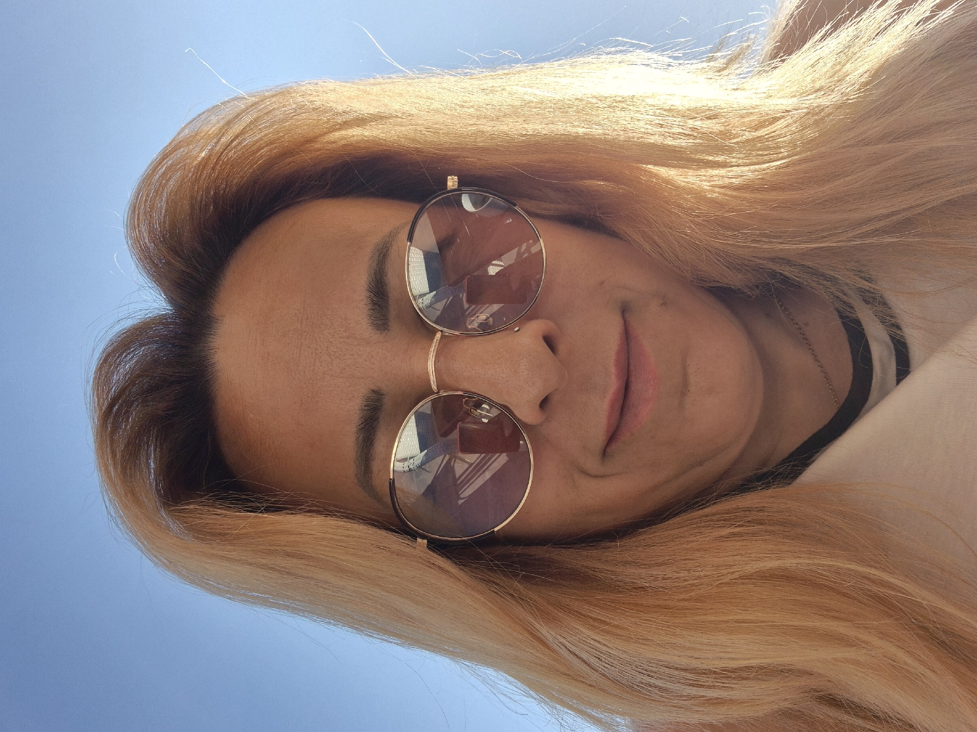 A close-up, low-angle selfie shows a person looking upwards against a clear blue sky. The person has long, blonde hair that is styled in loose waves and is illuminated by sunlight. They are wearing round, gold-rimmed sunglasses that reflect the surroundings, showing a glimpse of what appears to be a room with furniture and a phone. The person's facial features are partially visible, with closed eyes and a slight smile. A faint necklace is visible on their neck, and they are wearing a shirt with black and white stripes. The lighting suggests it is daytime, likely outdoors due to the bright sky and the reflection in the sunglasses. The overall mood is relaxed and carefree.